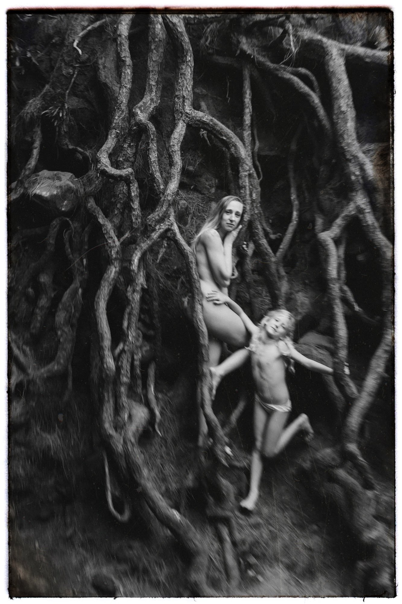 Black and white photo of a woman and a young child among large tangled tree roots, both partially unclothed.