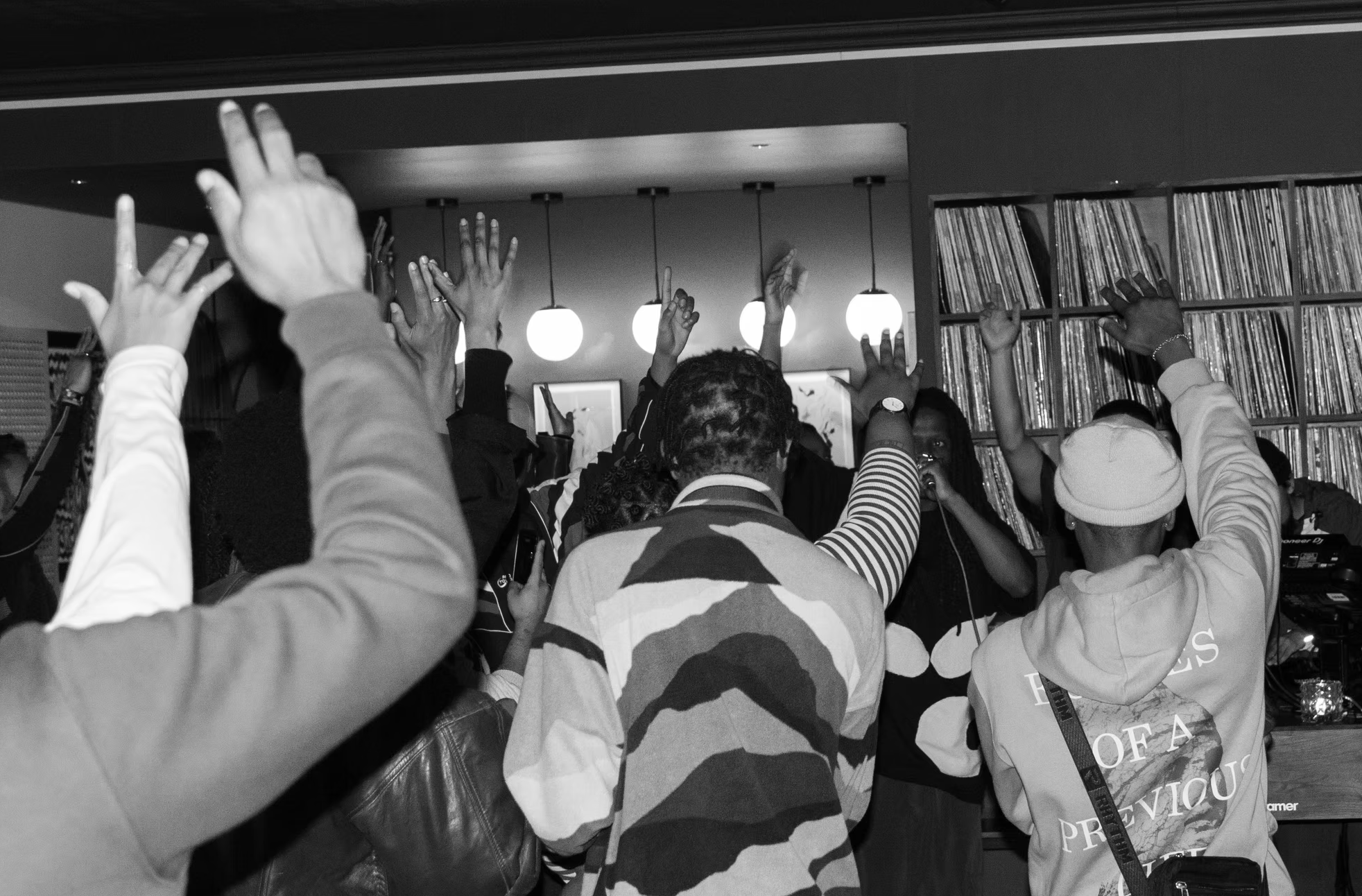 A group of people raising their hands during a gathering or event, with a woman holding a microphone in the background, in a room with art on the walls and shelves filled with records.