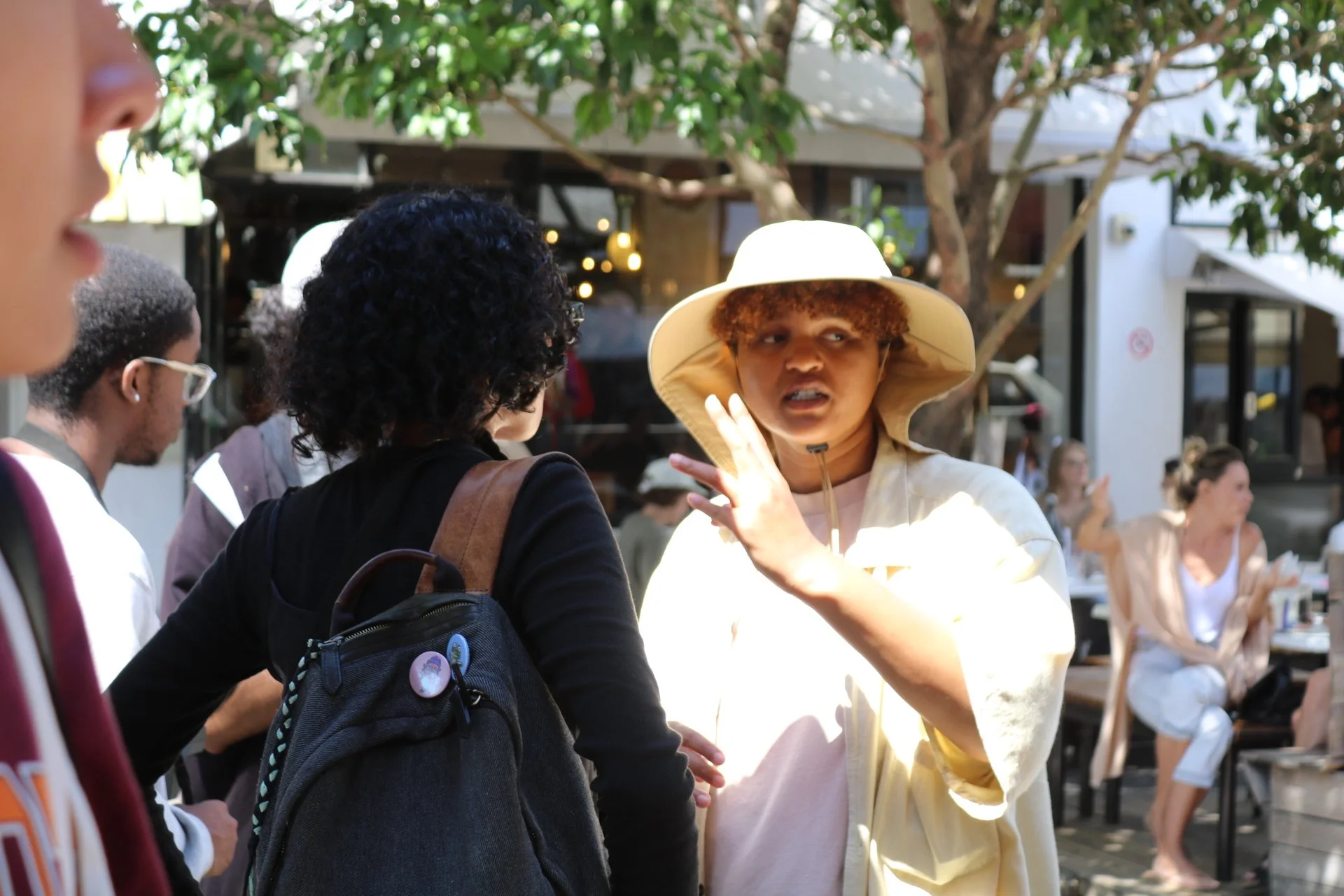 A woman in a wide-brimmed hat and light-colored clothing appears to be speaking to a group of people outdoors, with some sitting at tables in the background.