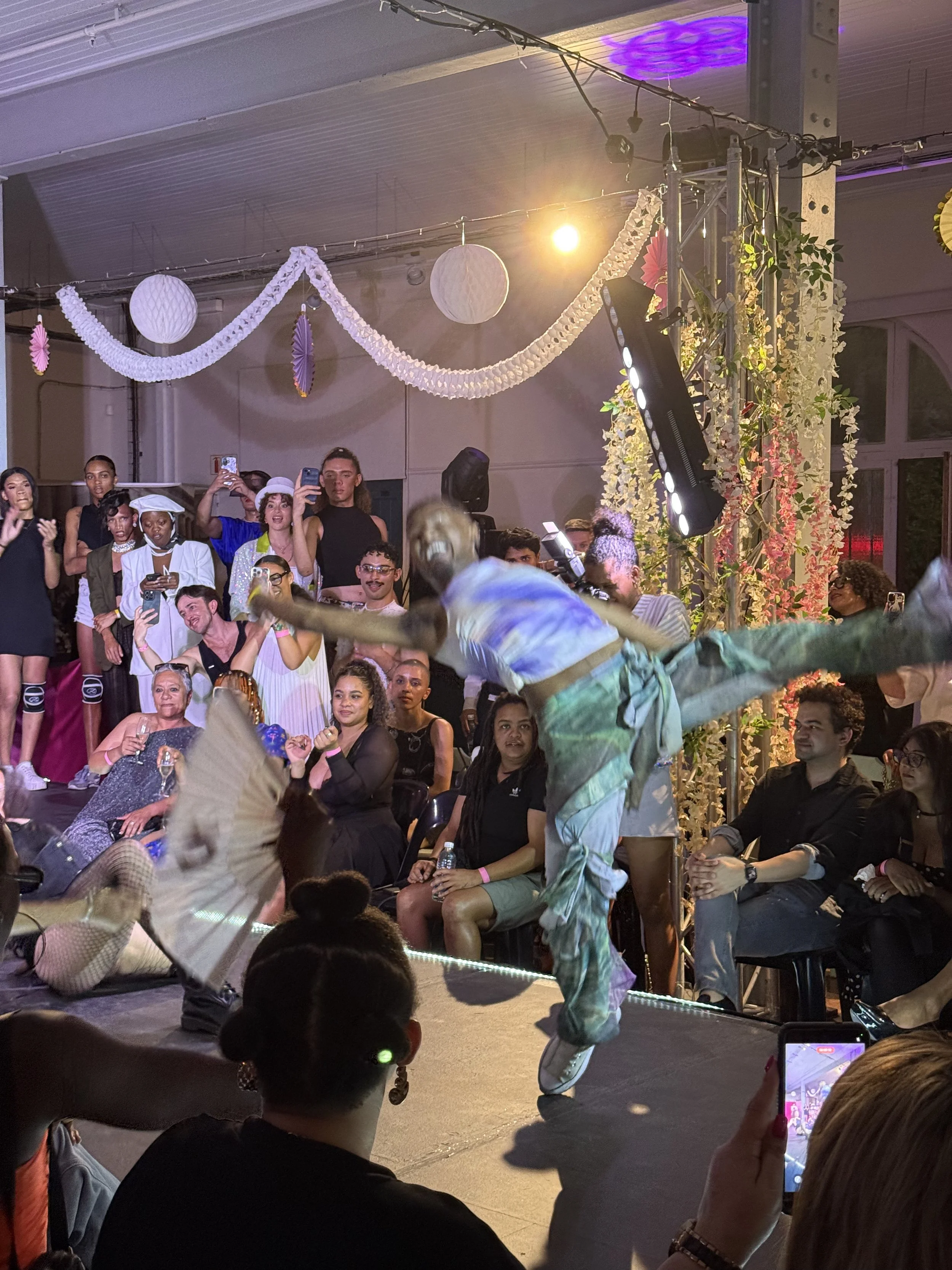 A performer in colorful clothing is mid-dance jump on stage at a lively event with an audience watching and recording. The stage is decorated with hanging paper lanterns and flowers, with lighting and sound equipment visible.