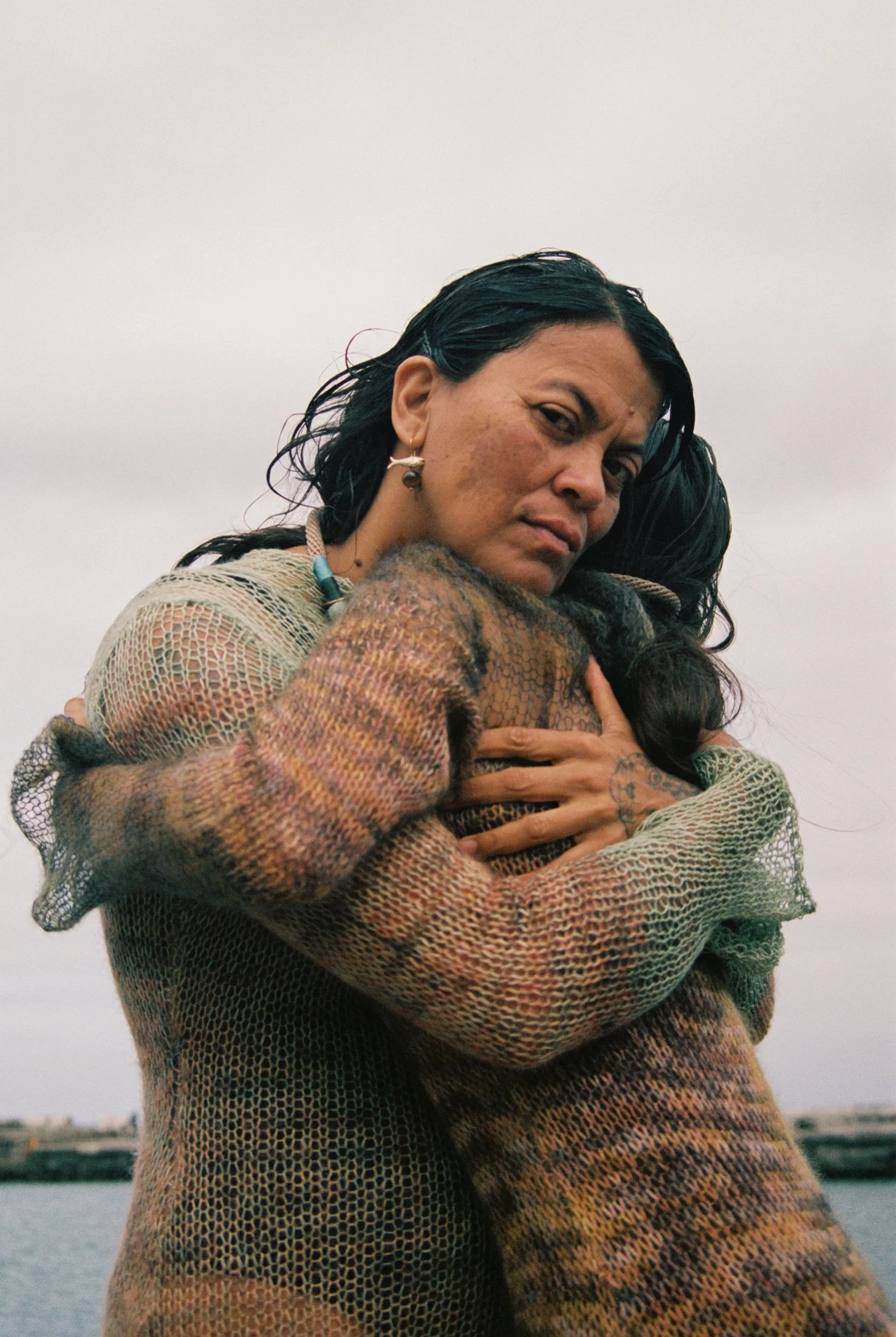 A woman with dark wavy hair embraces a person wearing a colorful, knit sweater outdoors against a cloudy sky.