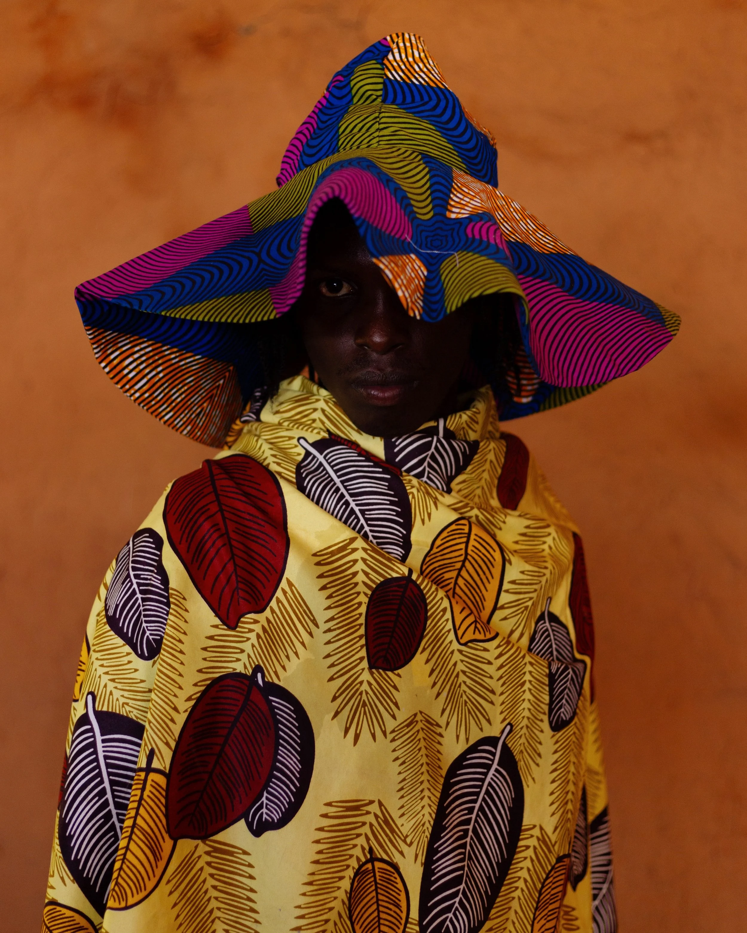 A person wearing a colorful, patterned hooded outfit that features large leaves in red, black, and gold on a yellow background. They are also wearing a wide-brimmed hat with vibrant blue, pink, orange, and green wavy lines, partially covering their face against a warm, blurred background.