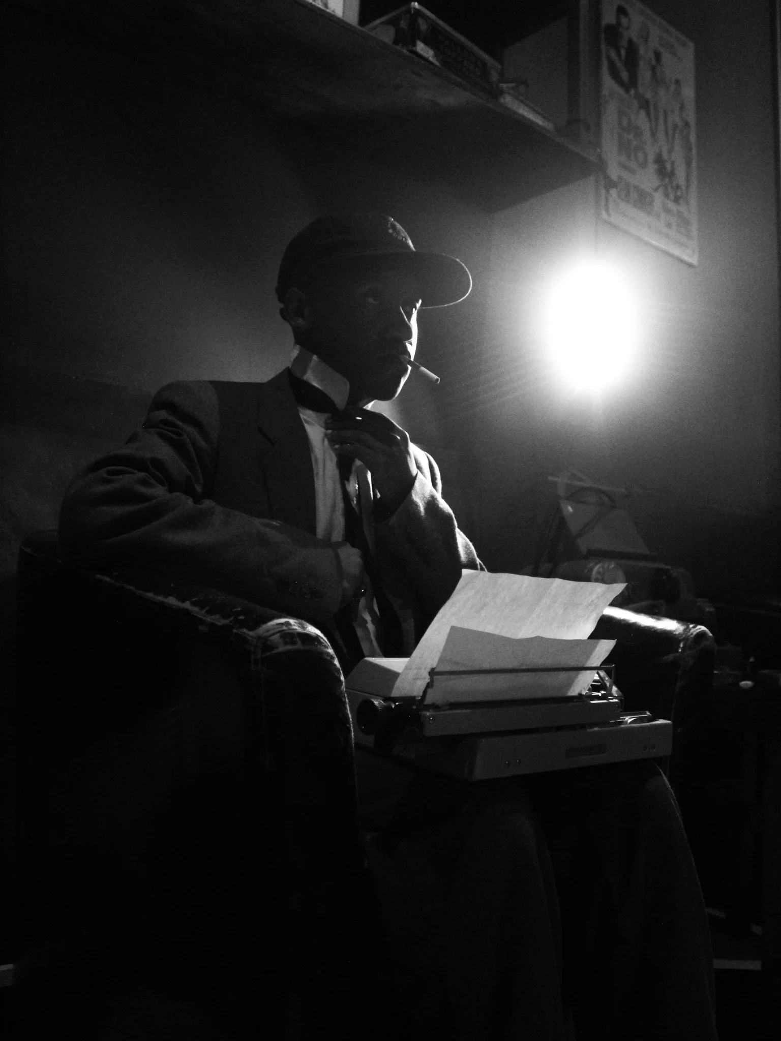 A black-and-white photo of a person sitting in a chair, wearing a hat and a suit, with a cigarette in their mouth. They are in a dimly lit room with a bright light behind their head, and there are papers and a typewriter in front of them.