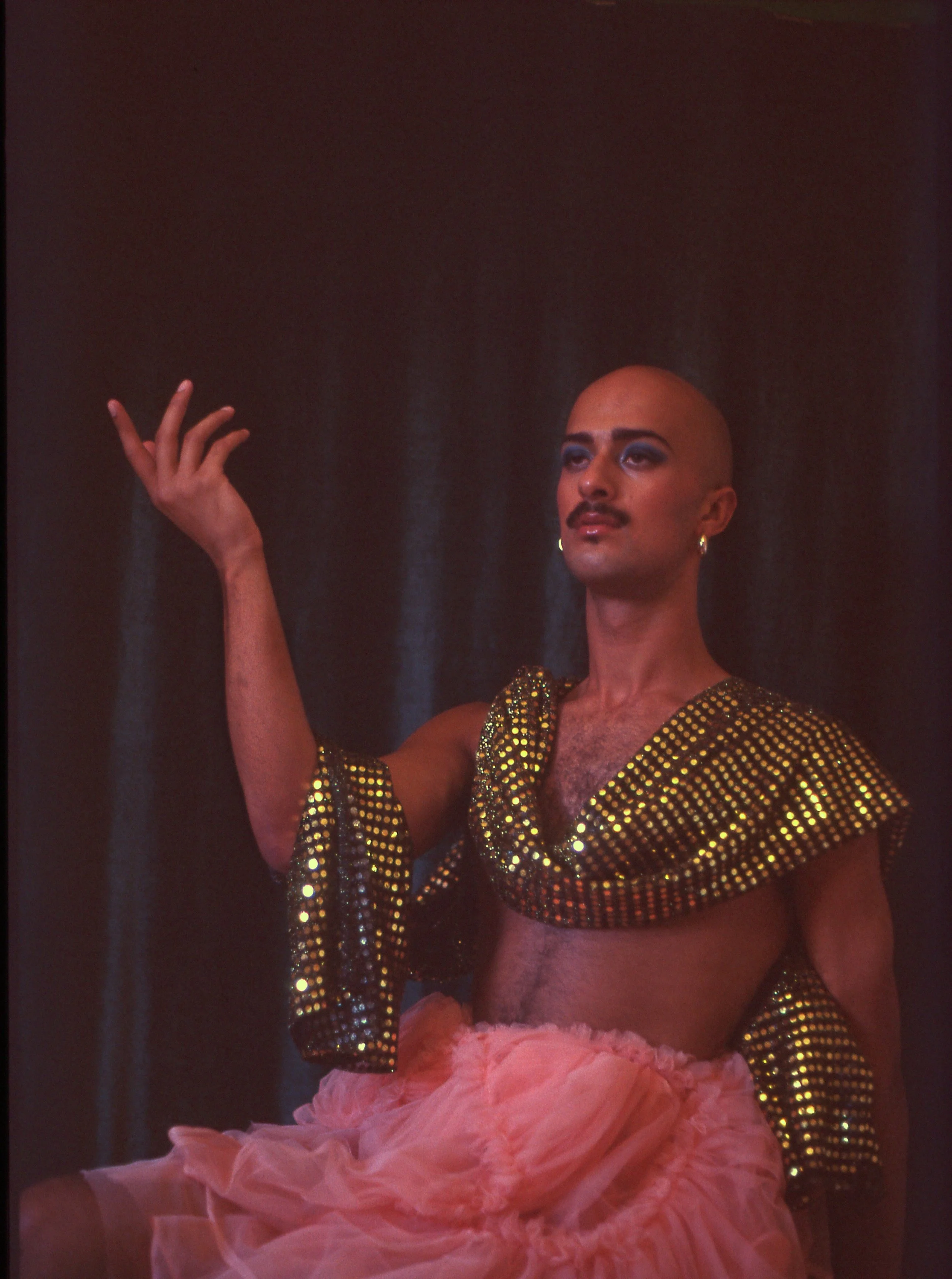 Person with a shaved head and bold makeup, wearing a shiny gold and black top with pink ruffles, posing in front of a dark background.