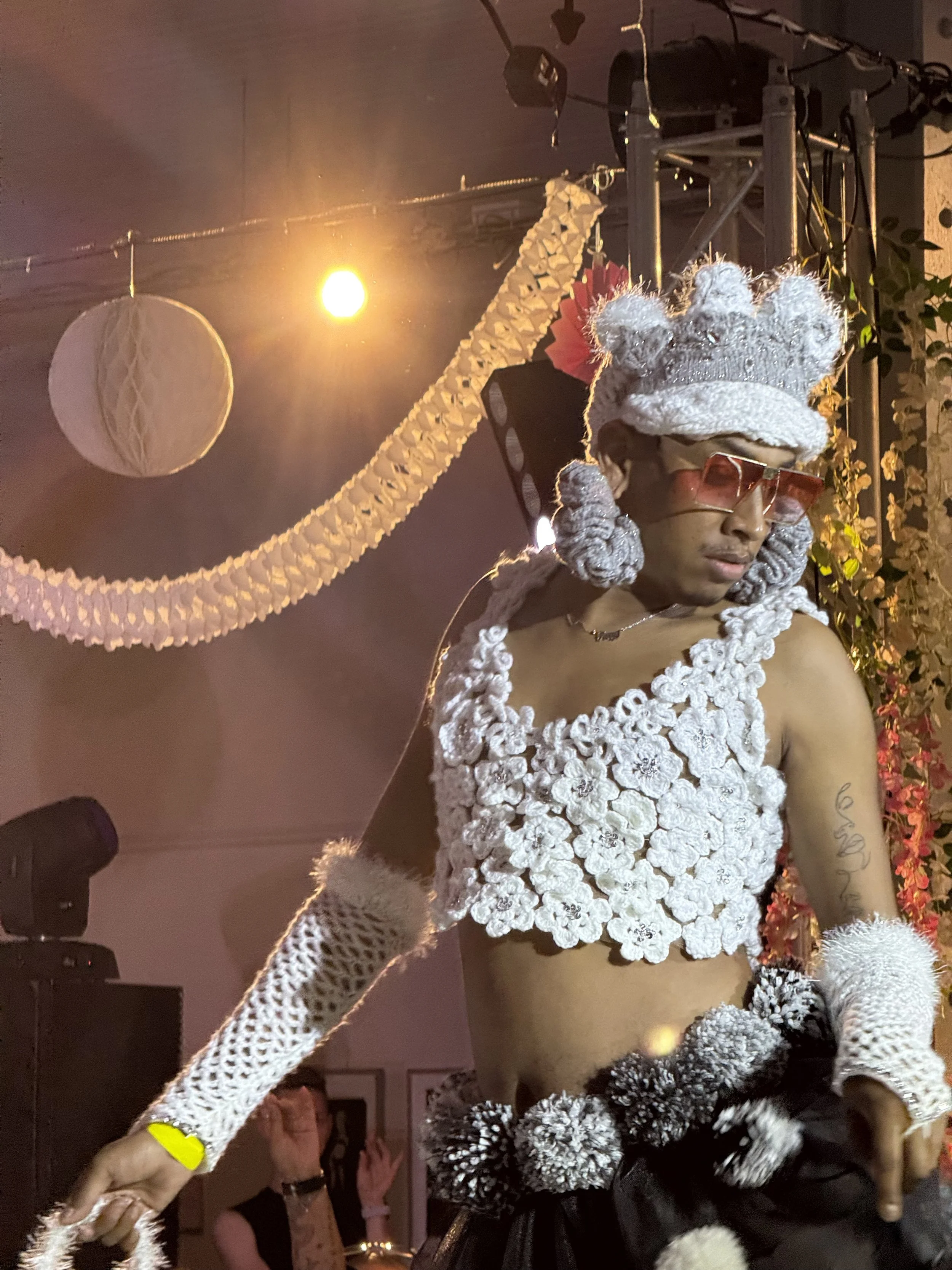 Person wearing a white, floral, crocheted top, furry accessories, pink-tinted sunglasses, and a large, decorative hat, dancing at a party or celebration with hanging paper decorations in the background.