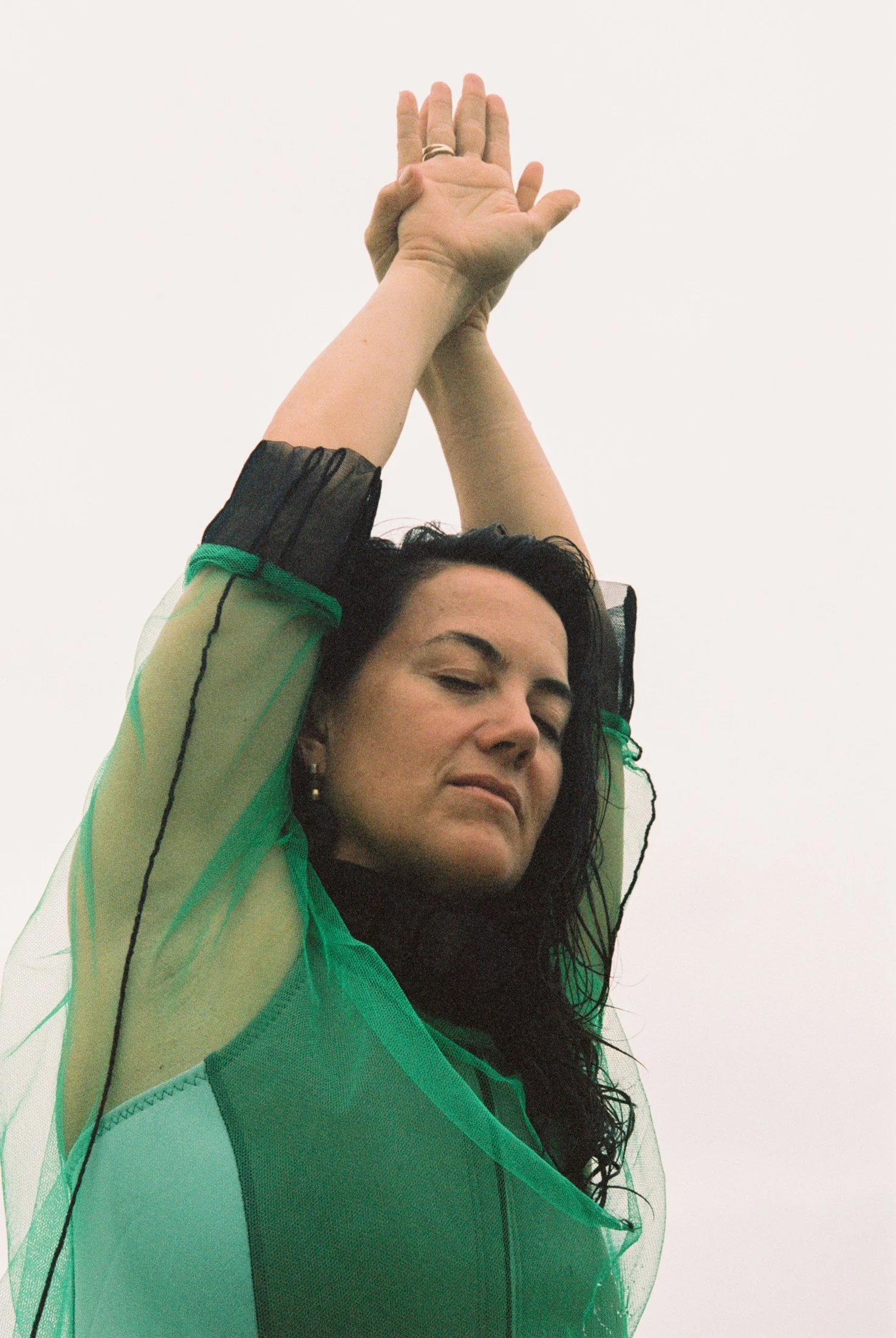 A woman with dark curly hair stretching with her eyes closed, wearing a sheer green and black top, against a plain white background.