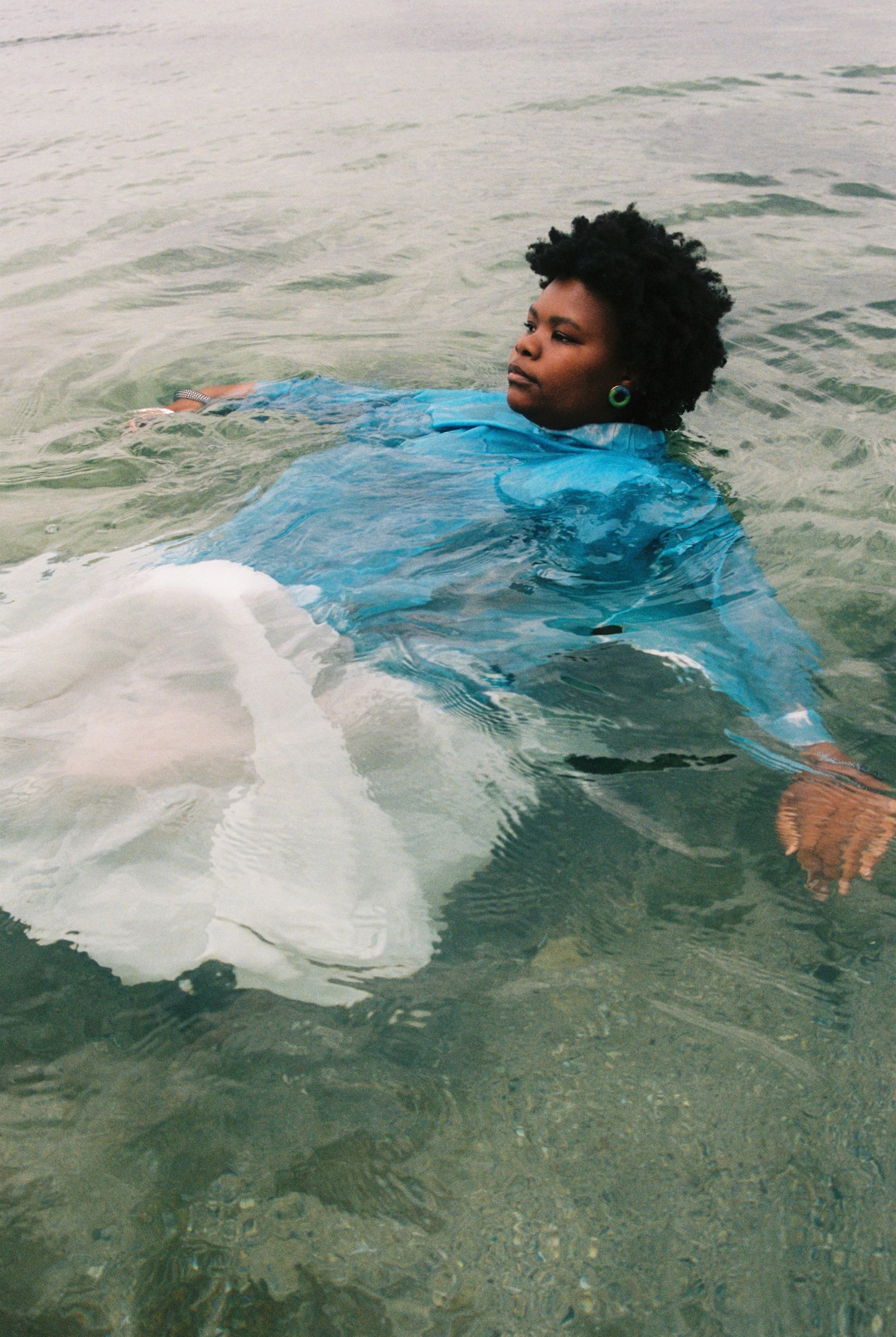 A woman with natural curly hair, wearing a blue shirt and green earrings, relaxing in clear shallow water at the beach with her arms outstretched.