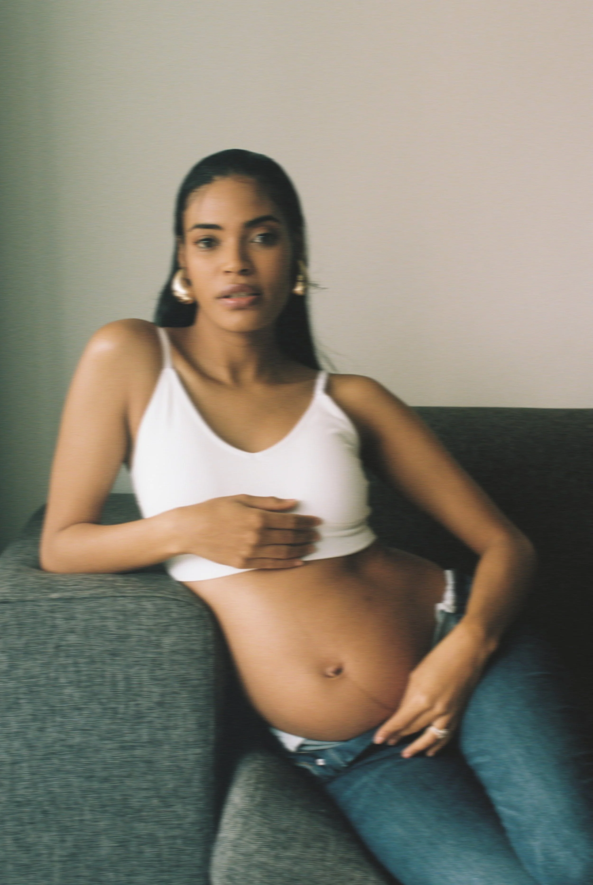 A pregnant woman sitting on a gray couch, wearing a white crop top and jeans, with her hand resting on her belly and her other hand on her chest, looking at the camera.