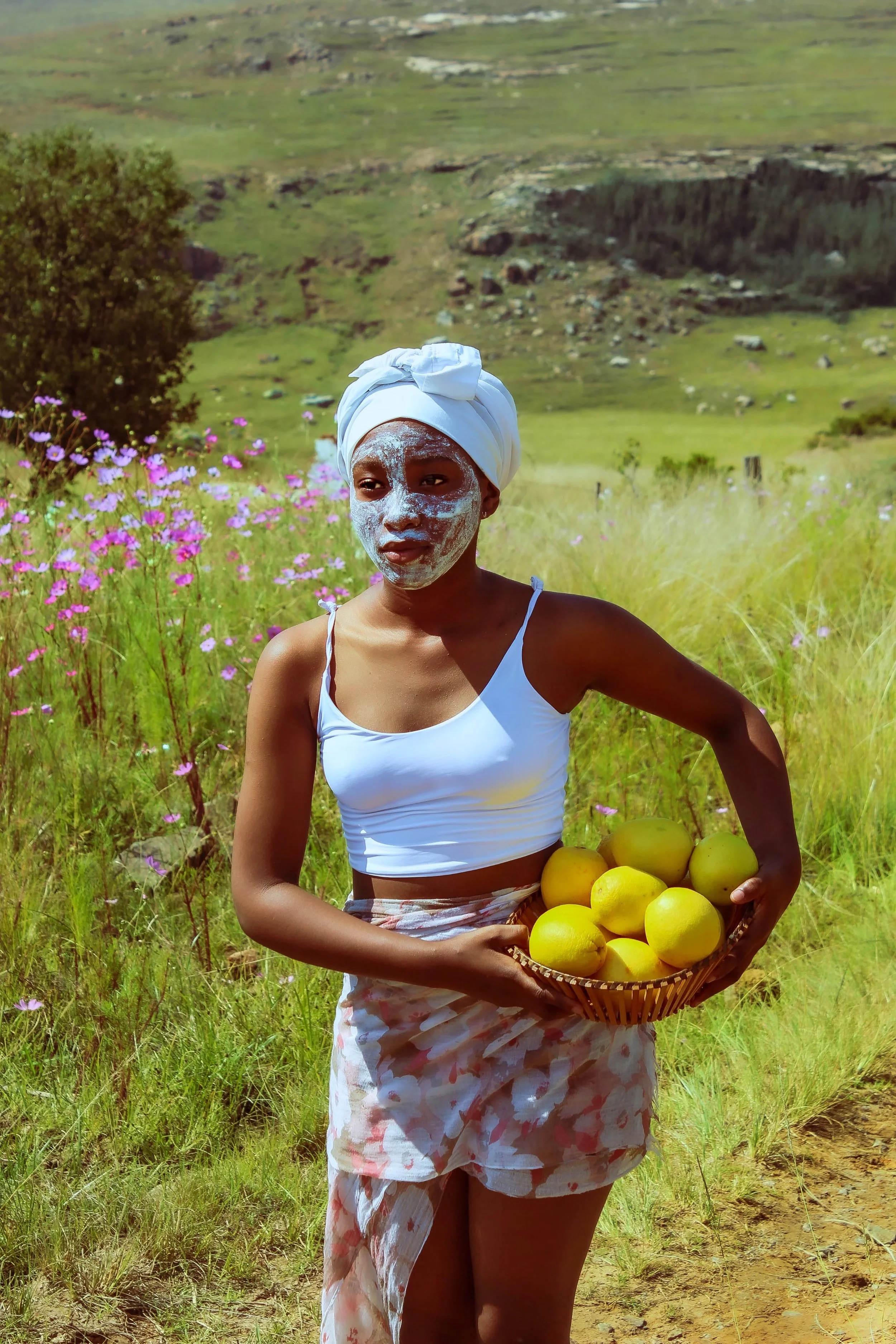 A woman standing outdoors in a rural field with wildflowers, holding a basket of yellow fruits, with hills in the background, wearing a head wrap, white top, and flower-patterned skirt, with facial mud.