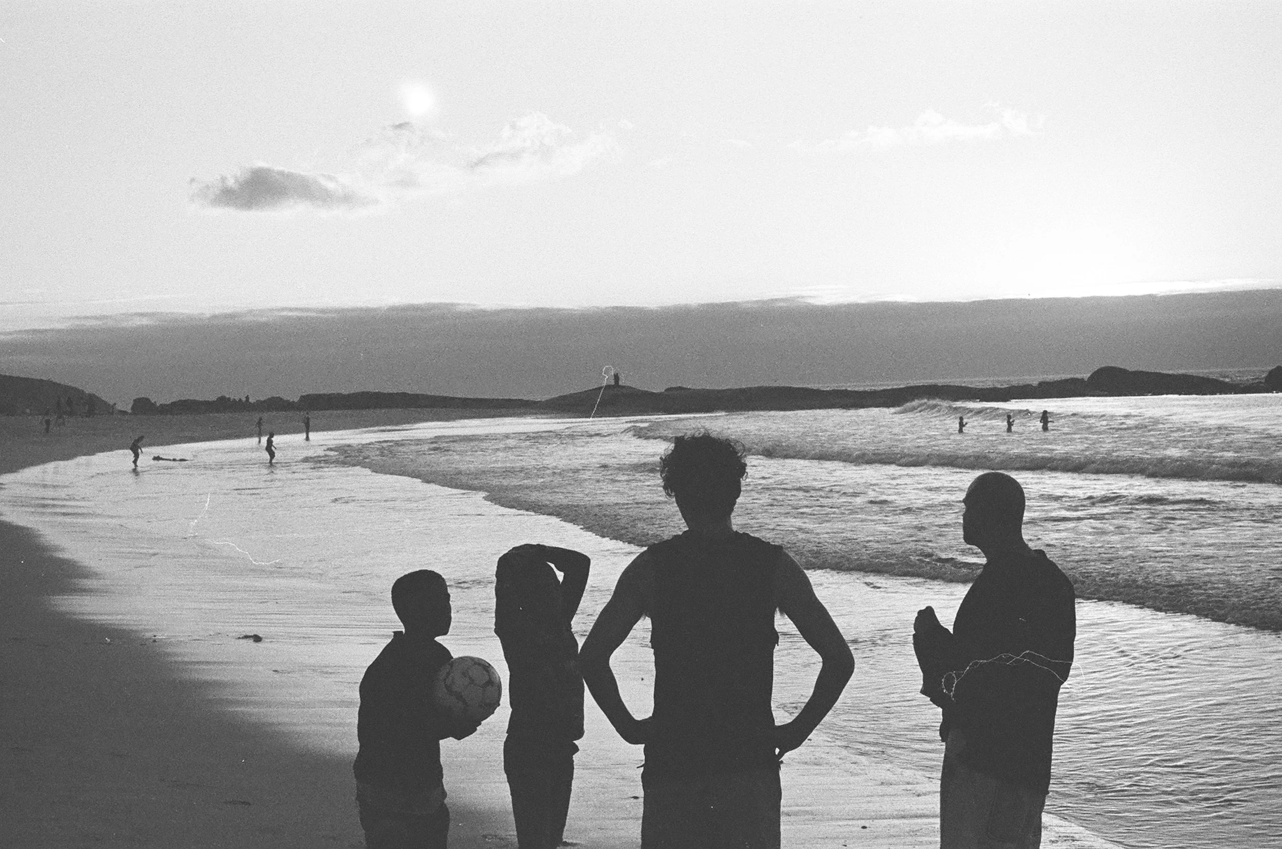 Silhouettes of four children on a beach with the ocean and other people swimming in the background, possibly during late afternoon or early evening.