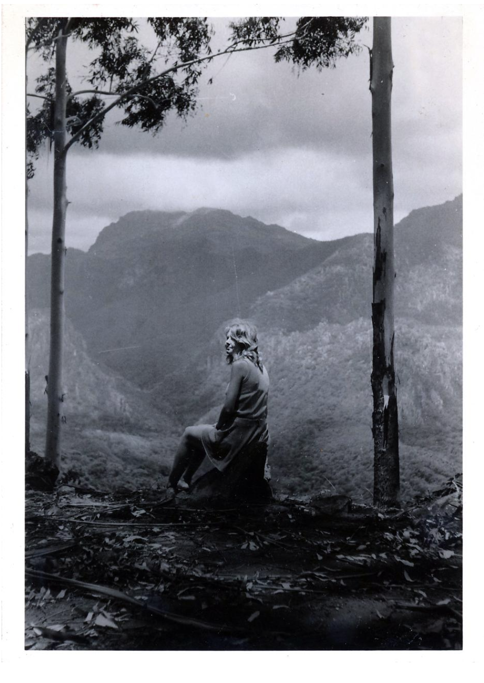 Black and white photo of a woman sitting on a rock in a mountainous landscape with trees and clouds in the background.