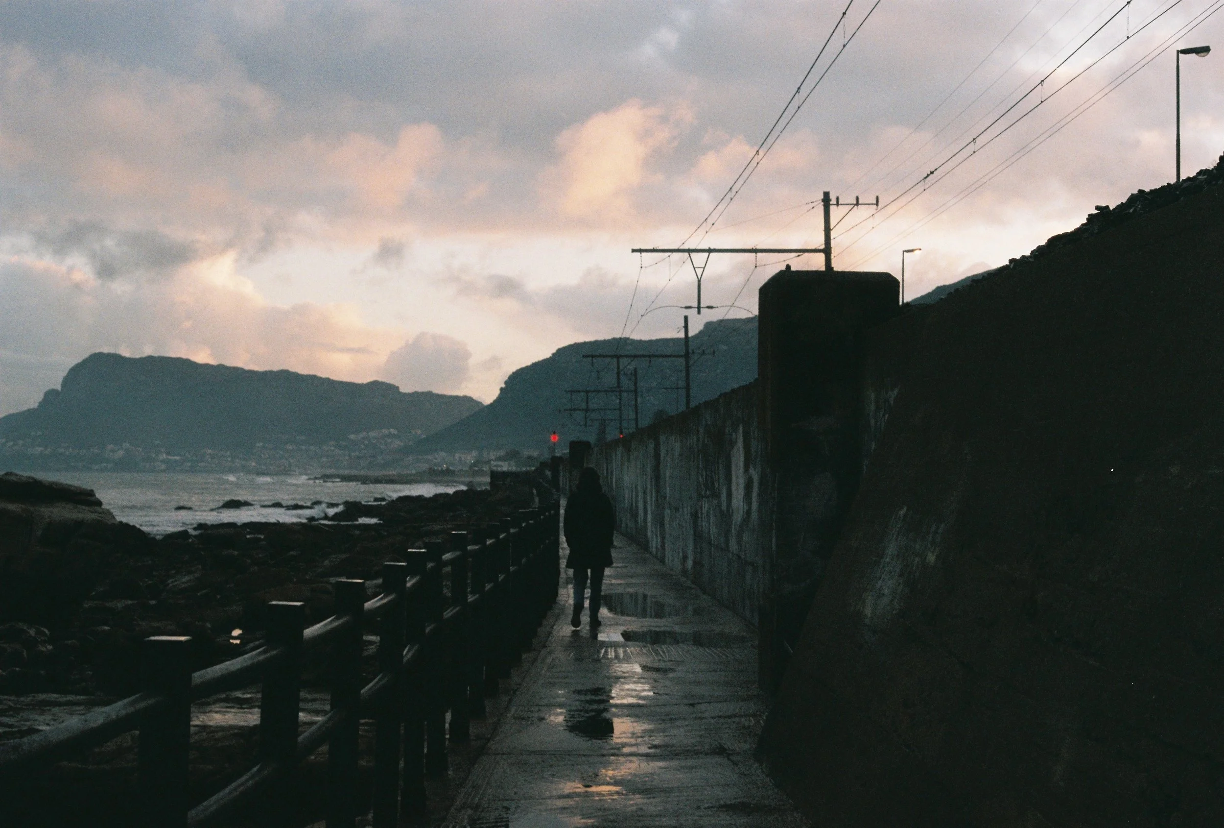 A person walking along a damp seaside walkway at dusk, with mountains in the background and power lines overhead.