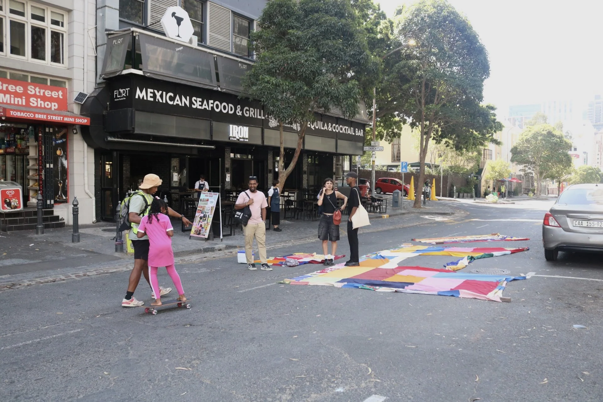 People gathered on a city street corner with colorful fabric laid out on the ground; a young girl skateboarding with an adult, three others chatting near a nearby restaurant.