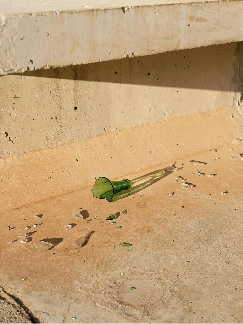 Broken green glass bottle on a beige sidewalk with shards of glass scattered around, against a brick wall.