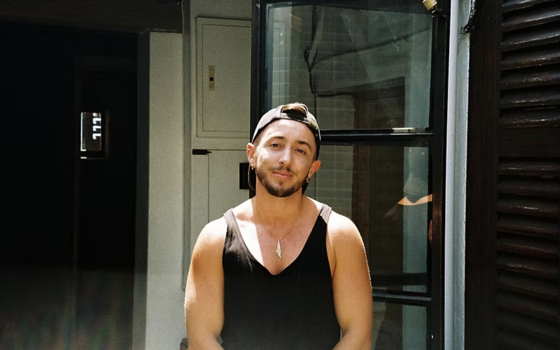 A young person with short dark hair, wearing a backward cap, earrings, and a sleeveless black shirt, standing outside near a window and smiling at the camera.