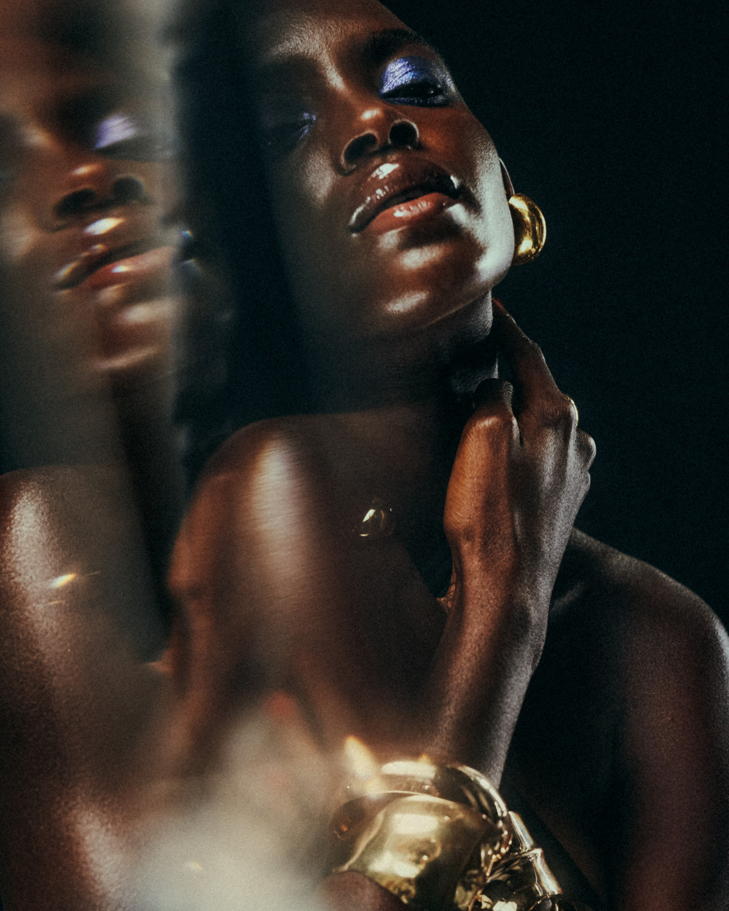 Close-up of an elegant woman with dark skin, wearing blue eyeshadow, glossy lips, gold earrings, a gold bracelet, and a hand touching her neck, with a blurred reflection of her face to her left.