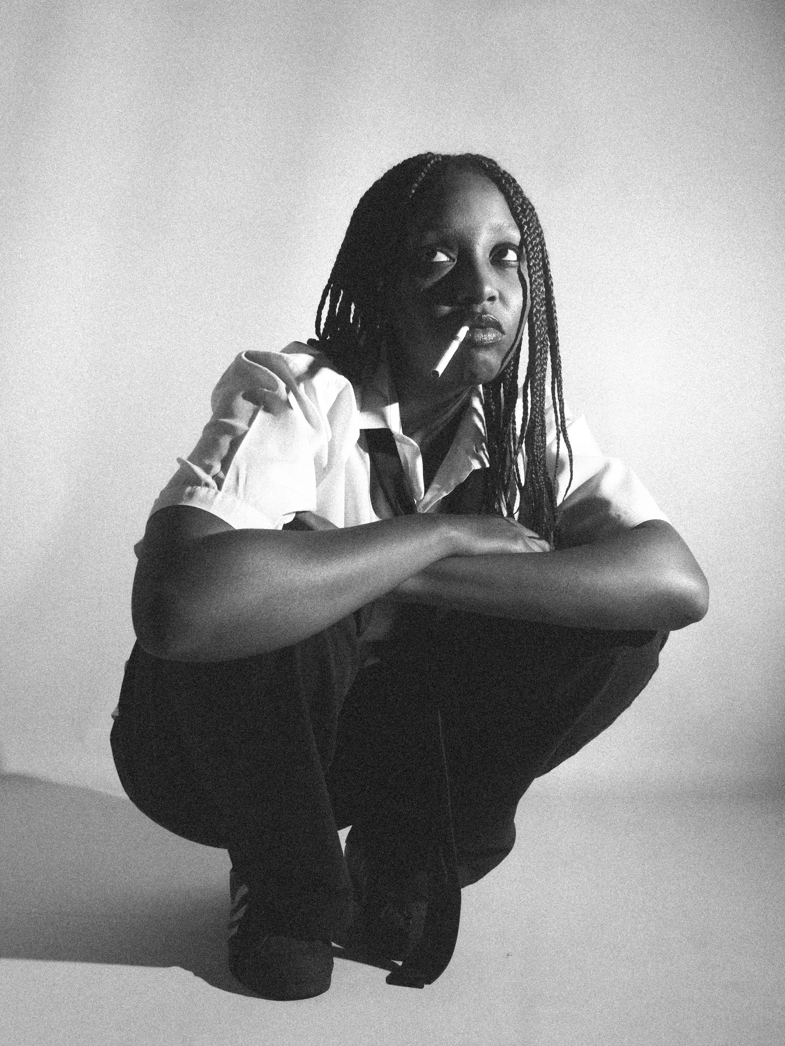 A woman with braided hair squatting against a plain wall, holding a cigarette in her mouth, wearing a short-sleeved shirt and dark pants.