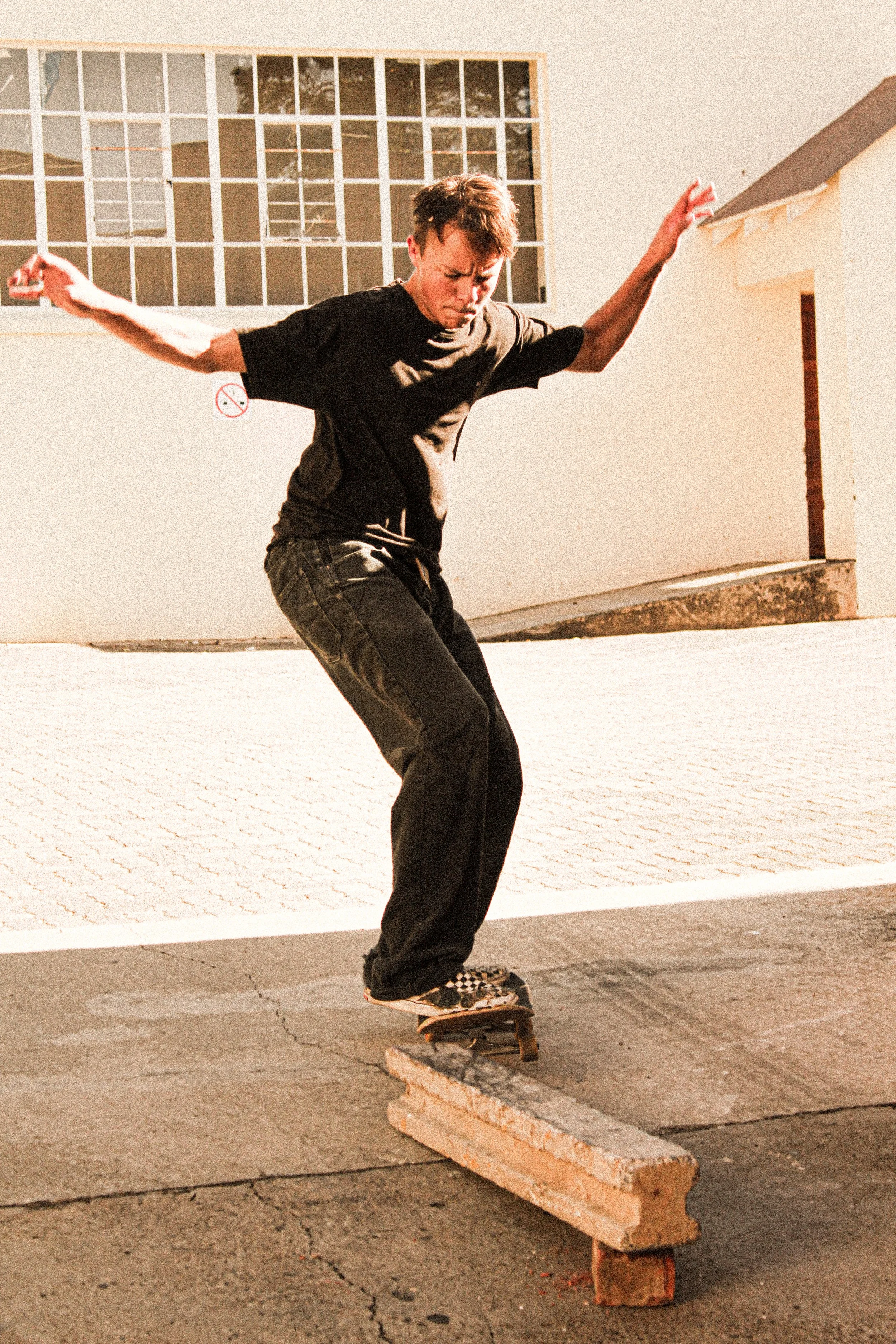 A young man wearing a black t-shirt, dark jeans, and checkered shoes is skateboarding over a wooden beam placed on the ground outdoors. The background shows a white building with large windows and a
