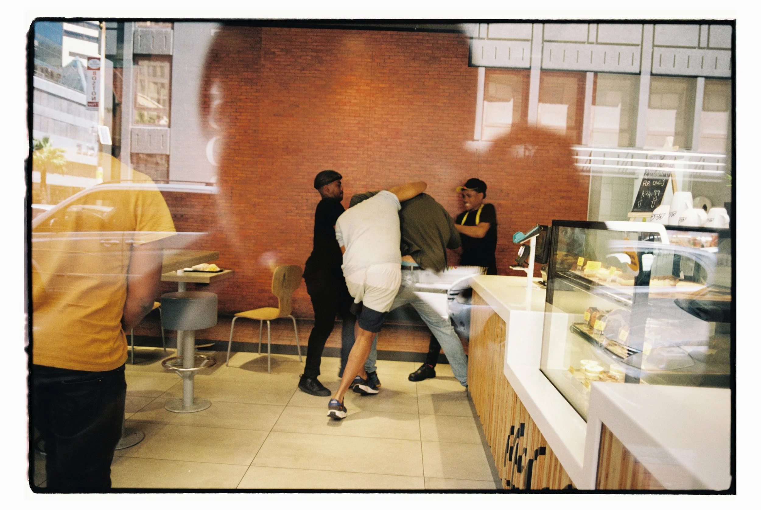 Several men fighting inside a cafe or bakery, with a man in a white shirt bent over a counter. The cafe has a glass display case with baked goods and a brick wall in the background.