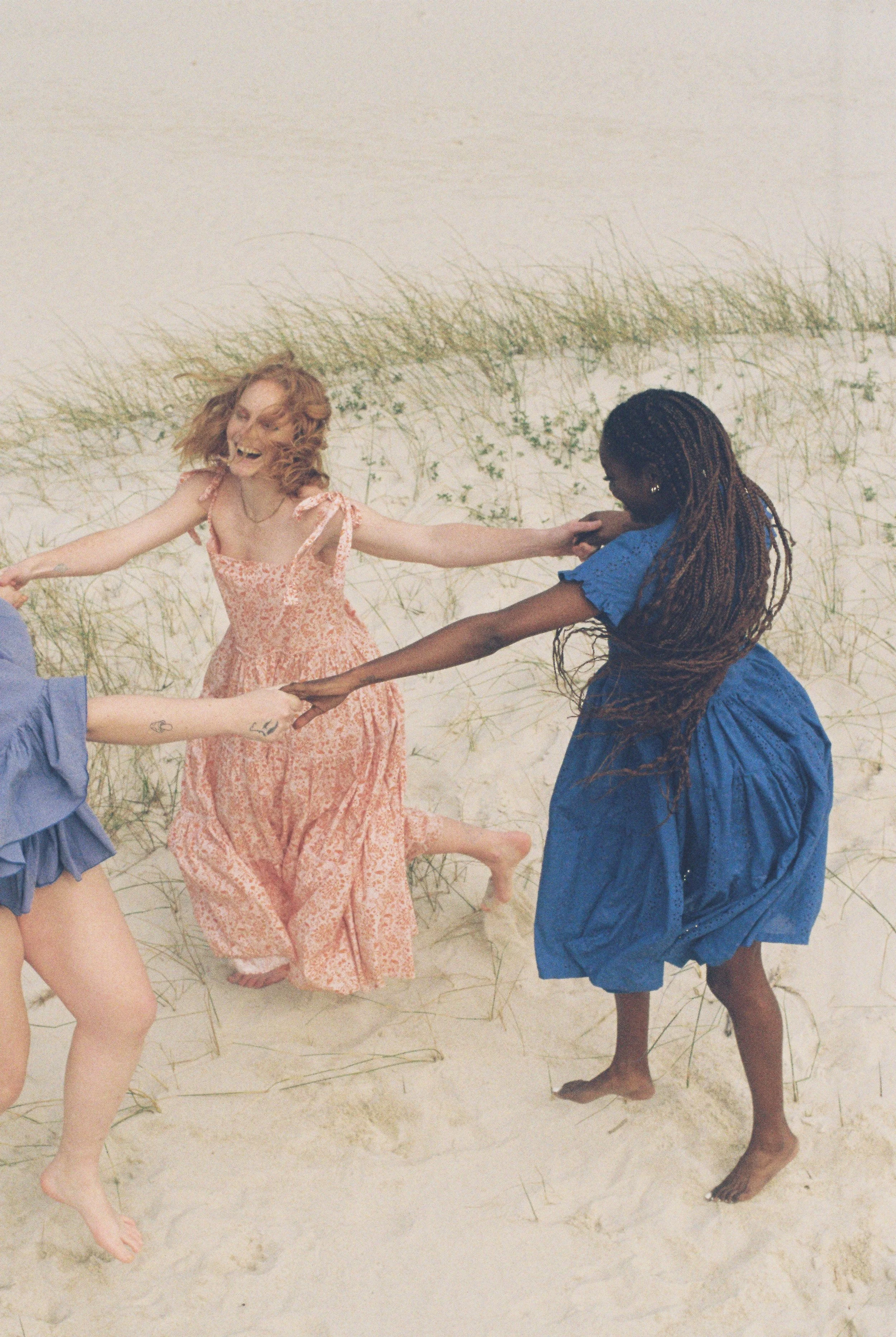 Three women holding hands and playing in a sandy beach area with grass in the background.