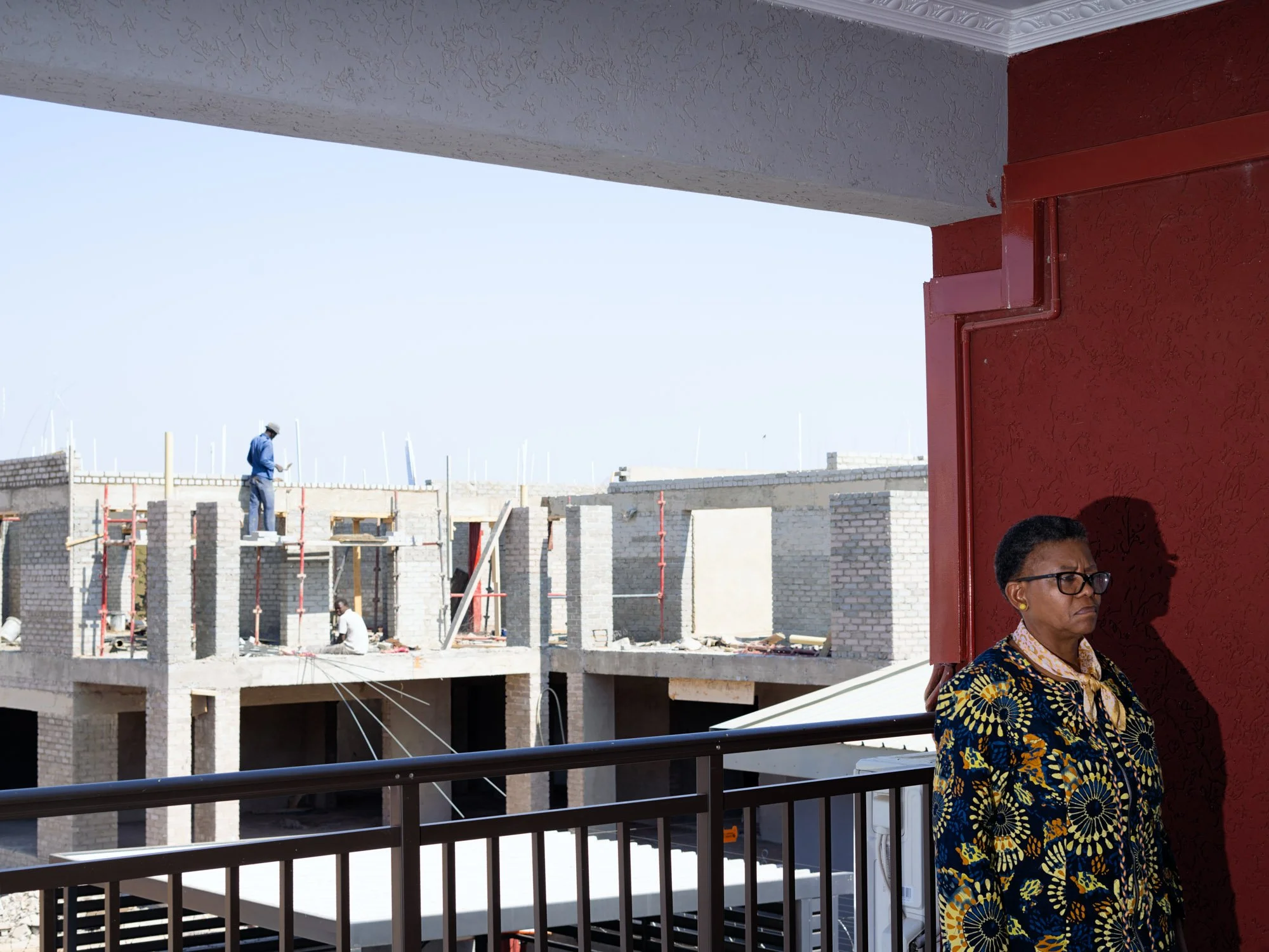 A woman standing against a red wall on a balcony, looking serious, with a construction site visible in the background.