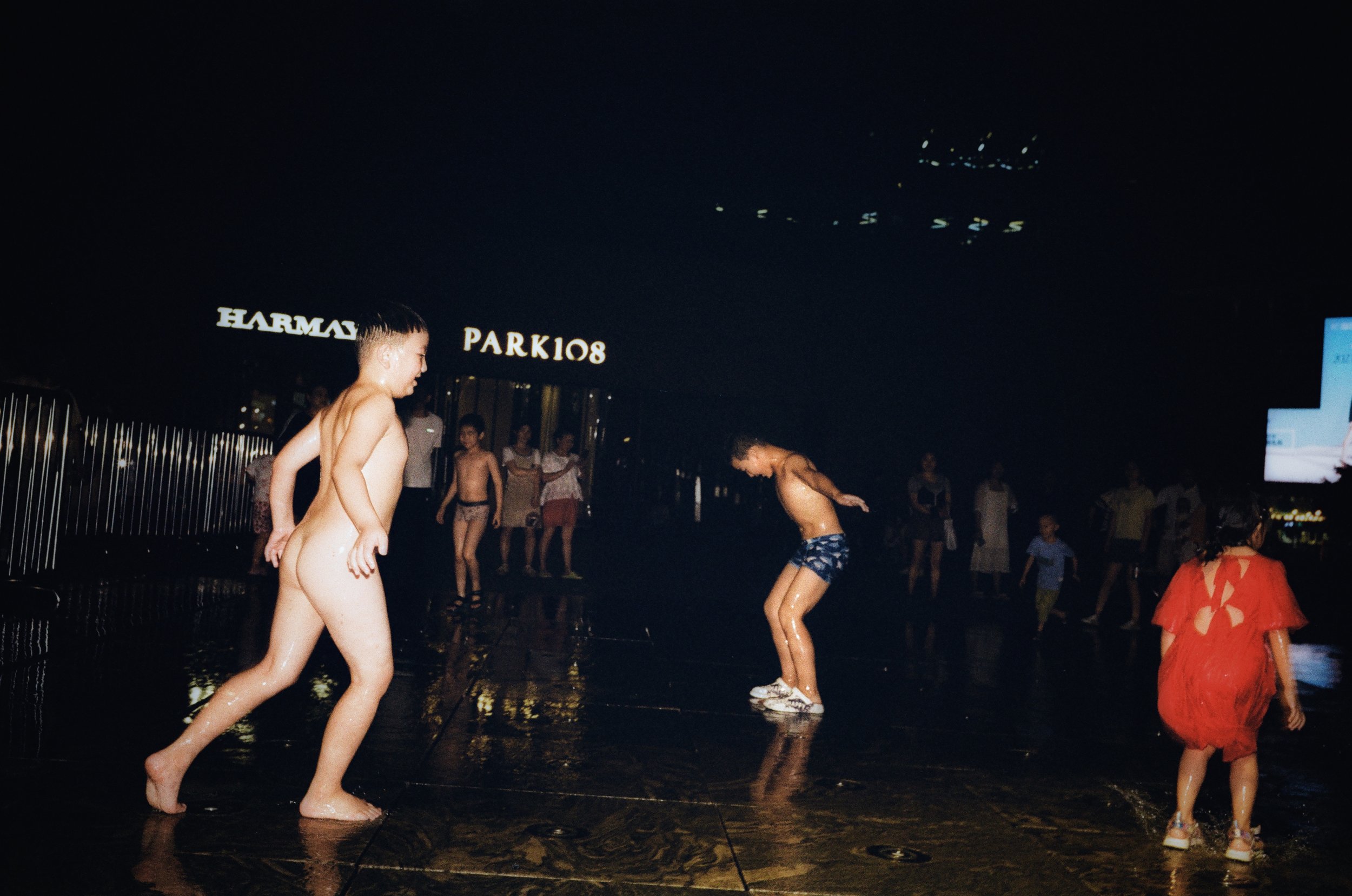 Children and adults playing and dancing in the rain on a wet outdoor surface at night. Some people are wearing swimsuits or light clothing, and there are illuminated signs and people in the background.