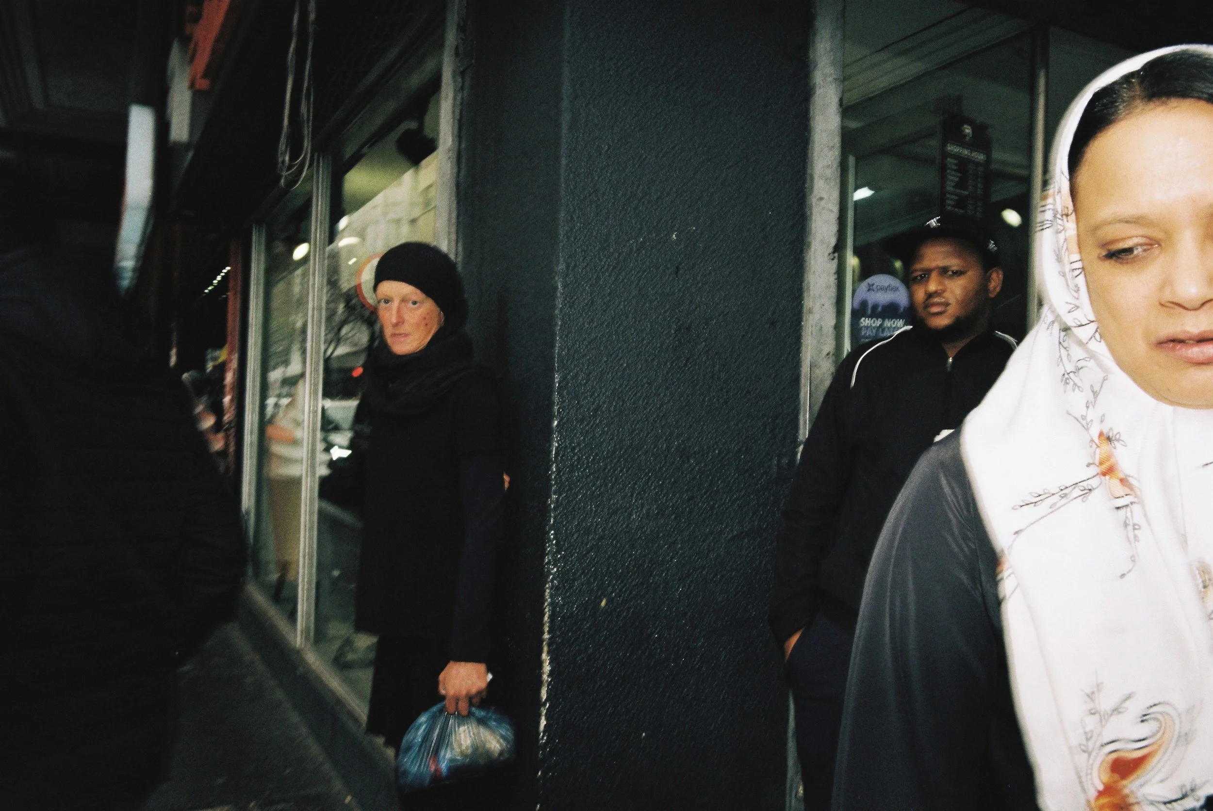 Three women and one man standing on a city sidewalk near a shop window, with a reflective glass and dark textured wall behind them, during what appears to be evening or night.