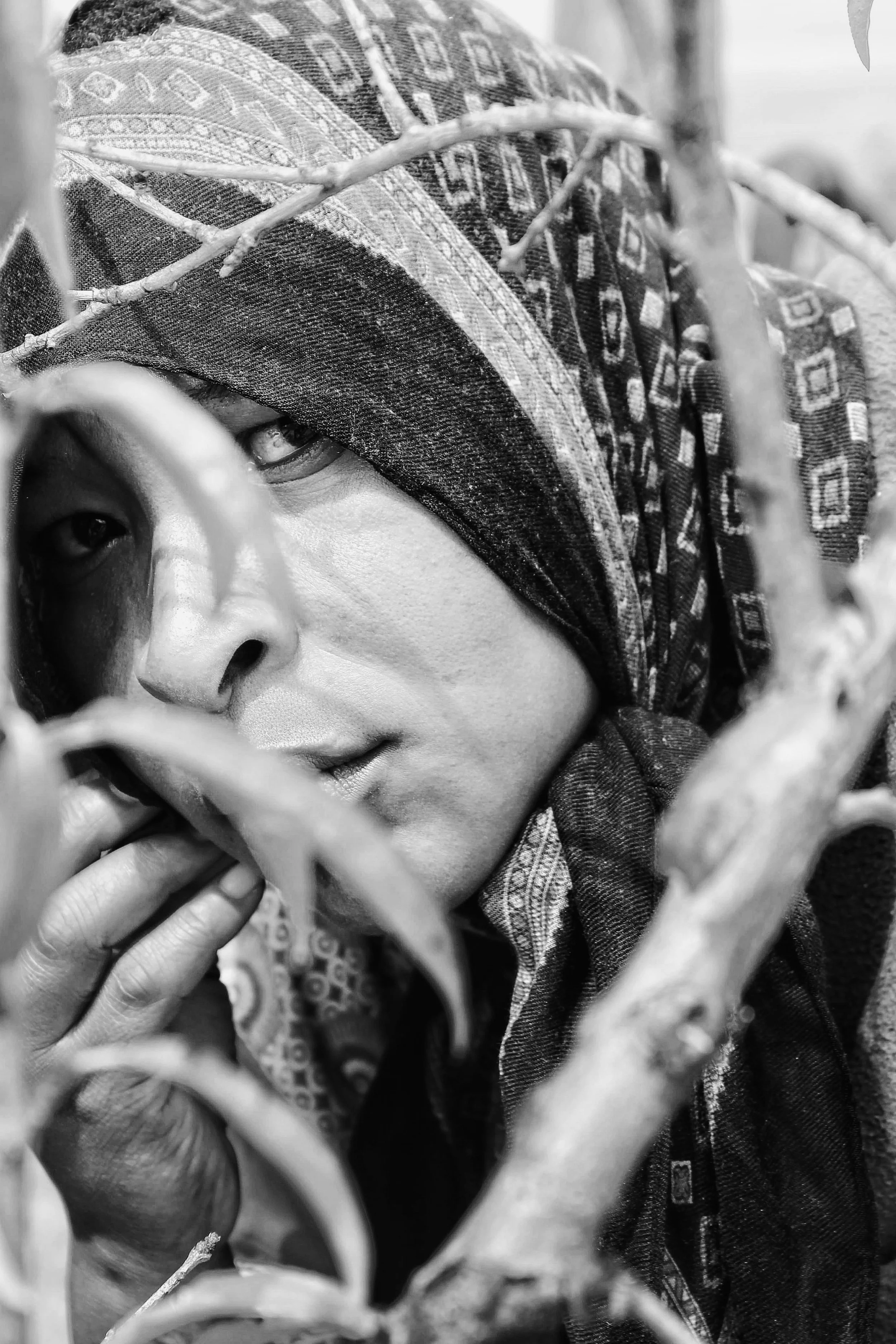 A close-up black and white photo of a person with a patterned headscarf, partially obscured by tree branches, gazing through the branches with a contemplative expression.