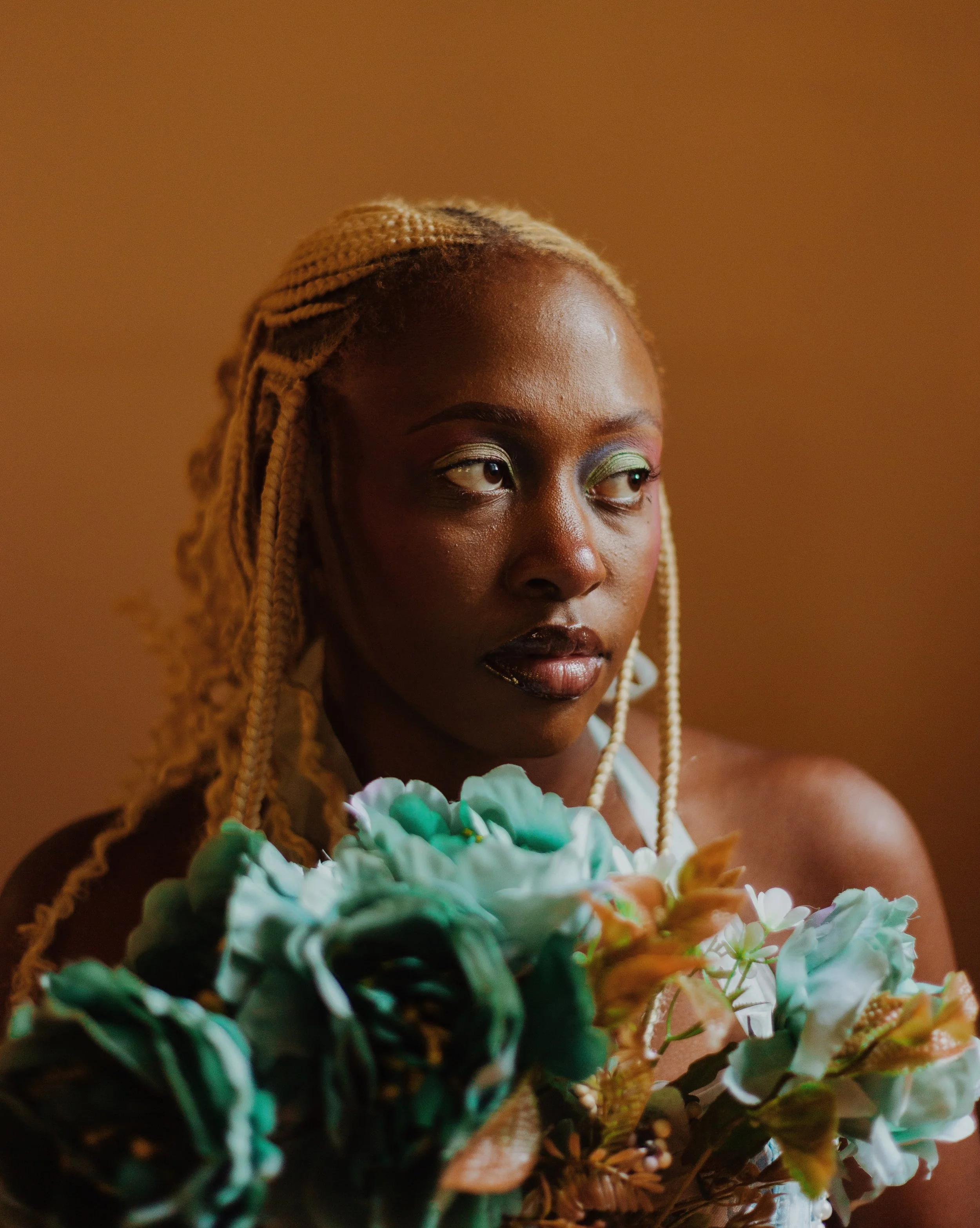 A woman with braided blonde hair and green eye makeup holding a bouquet of flowers, looking off to the side.