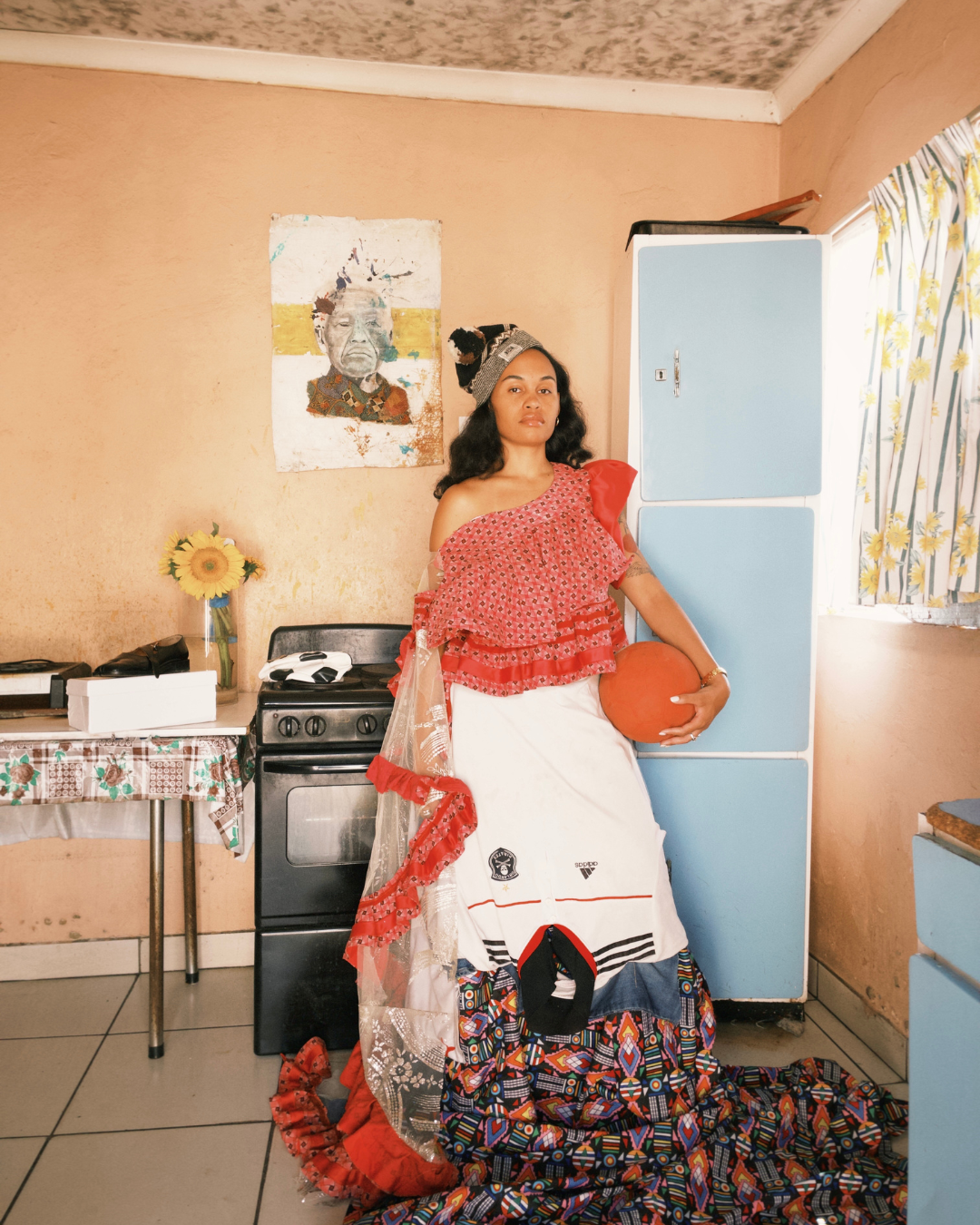 A woman standing in a kitchen, holding a basketball, wearing a colorful skirt, a layered, patterned top, and a beanie cap. Behind her, there is a blue refrigerator, a window with floral curtains, and a wall with a portrait of an elderly person. A small table with a sunflower in a vase and some items is beside her.