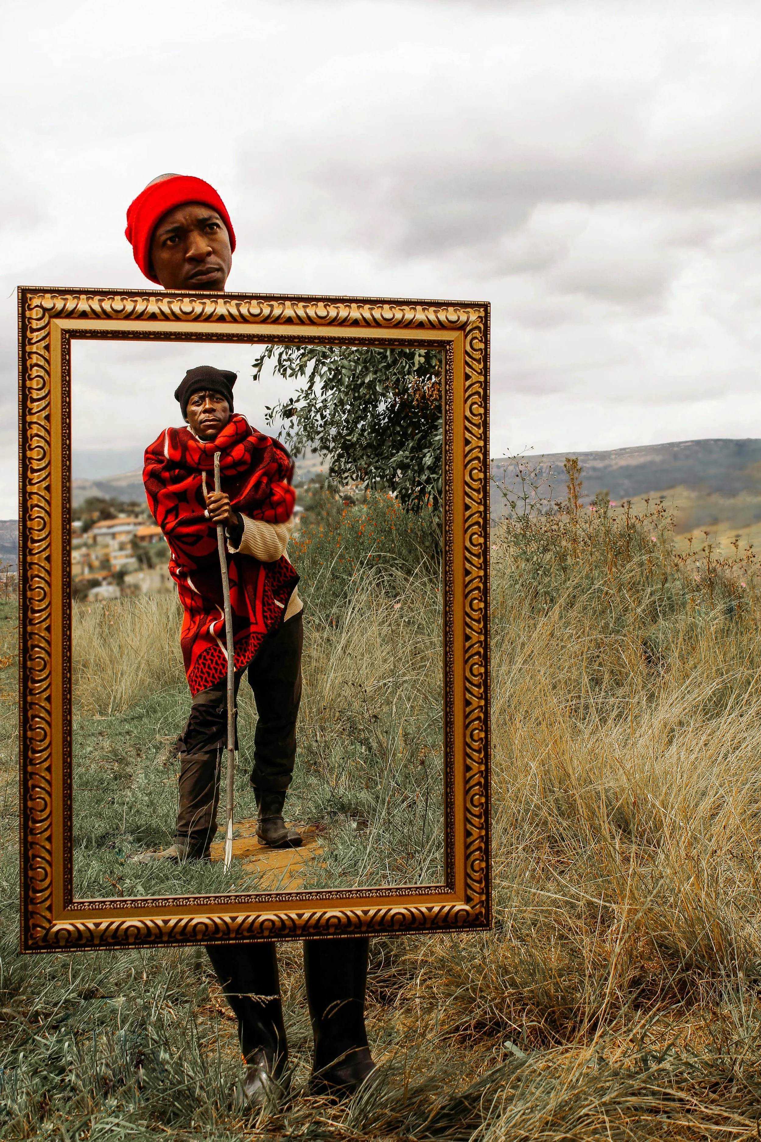 A person stands outdoors holding a large decorative mirror reflecting another person dressed in warm clothing with a scenic landscape and cloudy sky in the background.