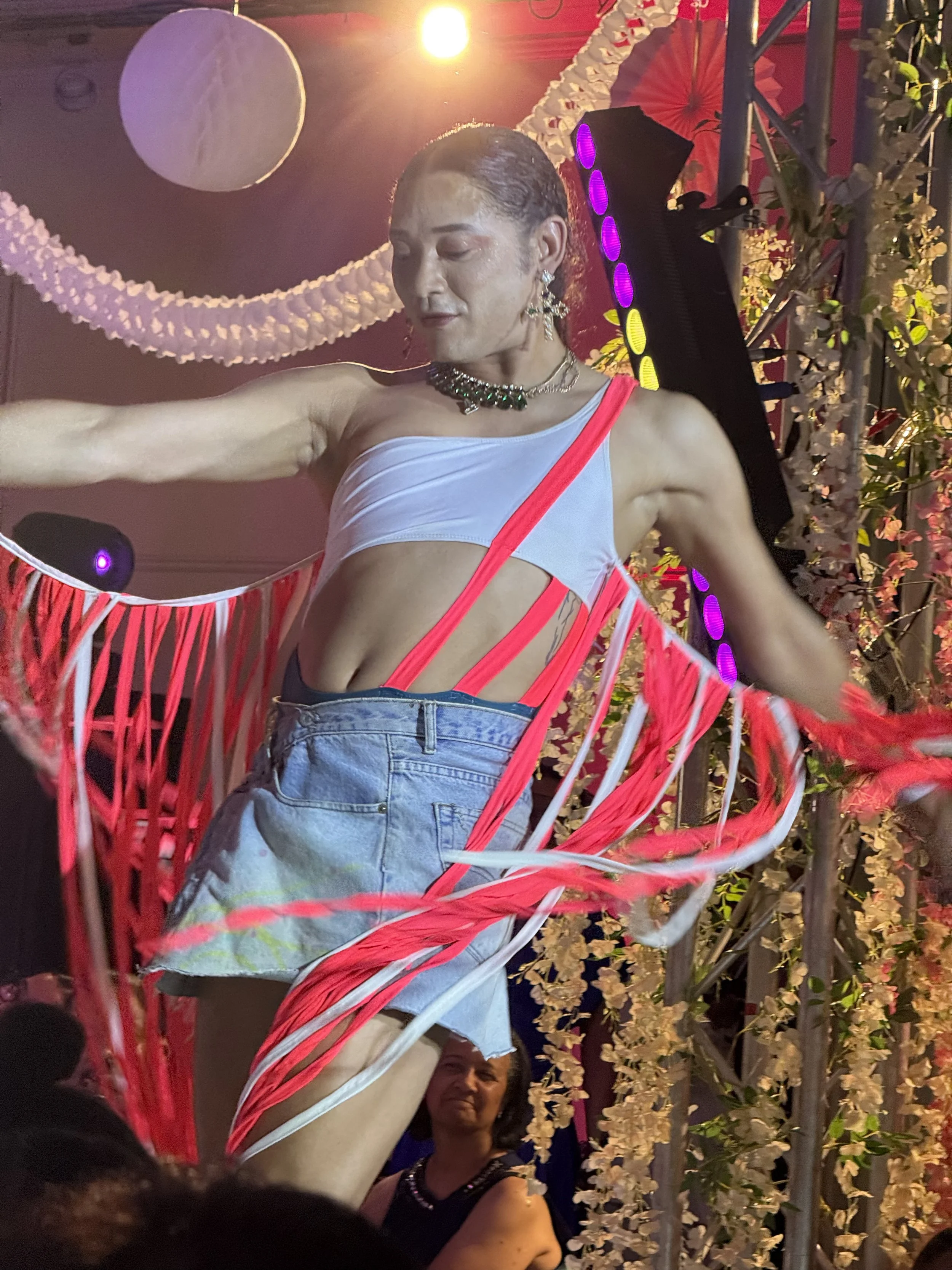 Person dancing on stage wearing a white and red costume with denim shorts, decorated with ribbons, jewelry, and earrings, possibly at a celebration or event with decorative lighting and floral arrangements.