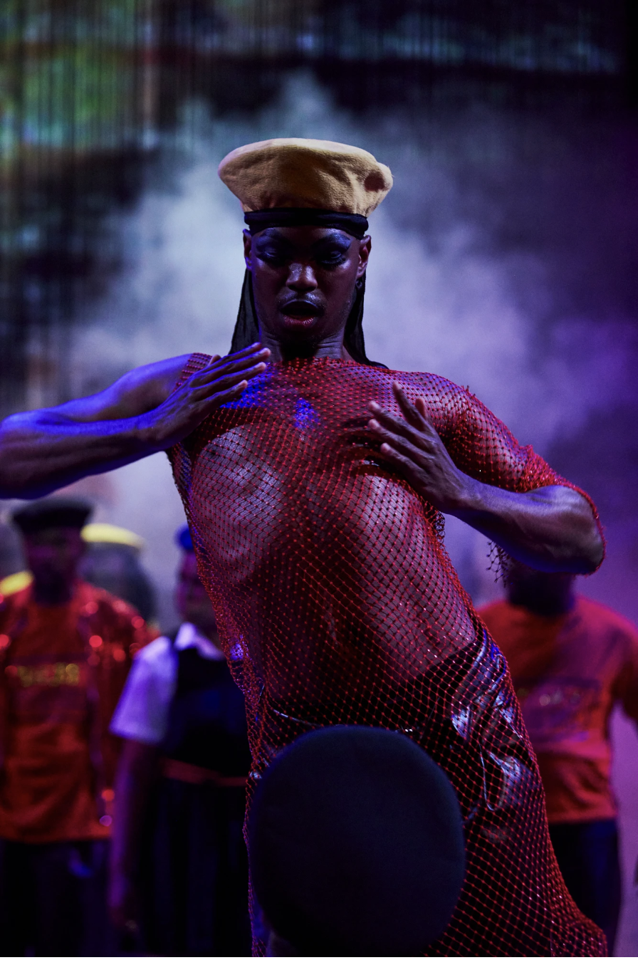 A performer wearing a beige beret, black headband, and red mesh dress on stage, surrounded by people in costumes, with colorful lighting and fog.