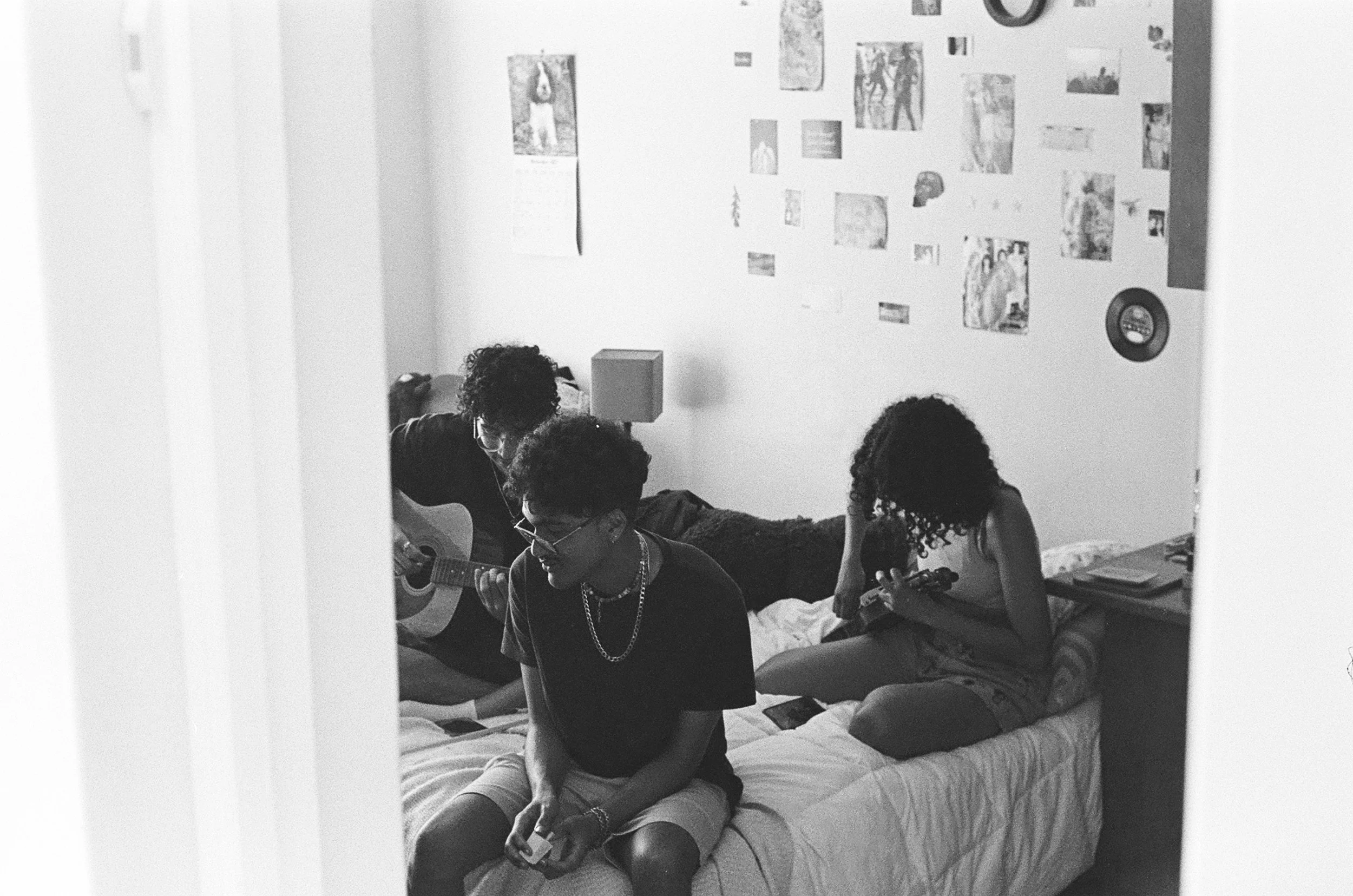 Three teenagers with curly hair in a bedroom, one playing guitar, one sitting and smiling, and a girl looking at her phone, in black and white.