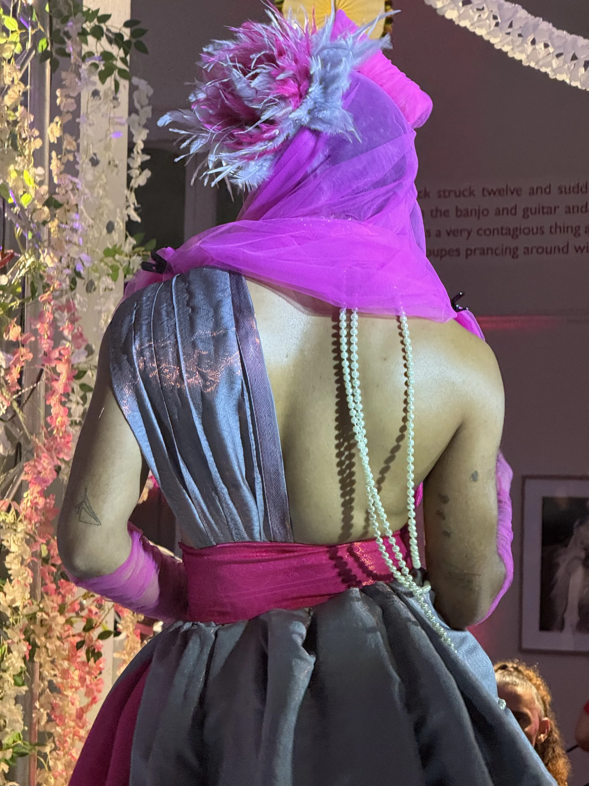 Woman with a pink headscarf decorated with feathers, wearing a strapless dress with grey and pink fabric, long pearl necklace, and pink gloves, viewed from behind in an indoor setting with floral decorations.