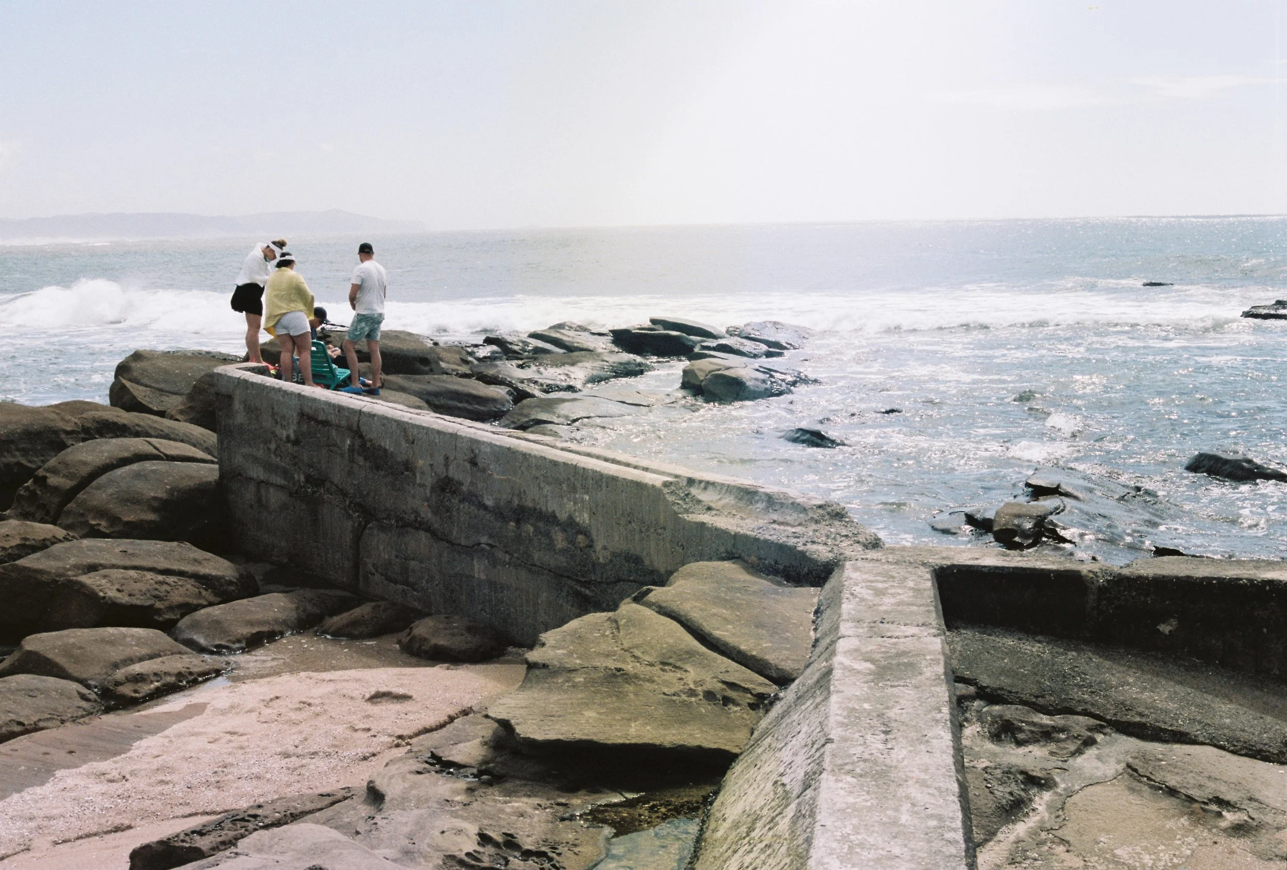 Group of people standing on concrete and rocky shoreline by the ocean, with waves and distant land in the background.