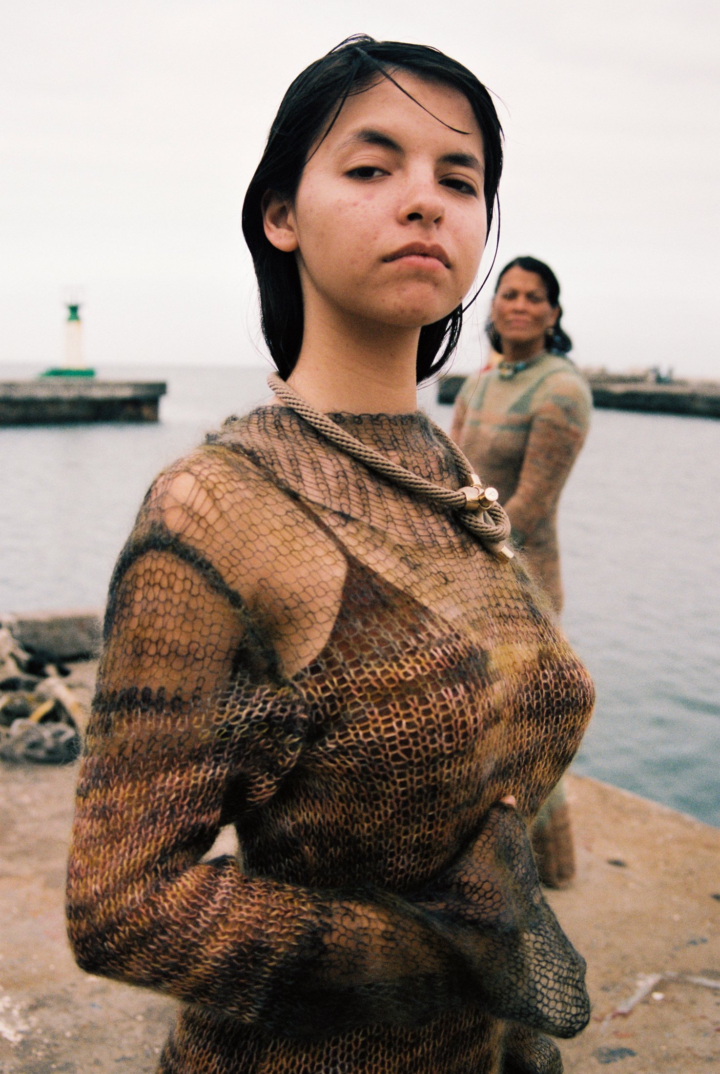 A young woman with short dark hair and a serious expression, wearing a see-through, patterned top, standing near a body of water with another woman in the background, overcast sky.