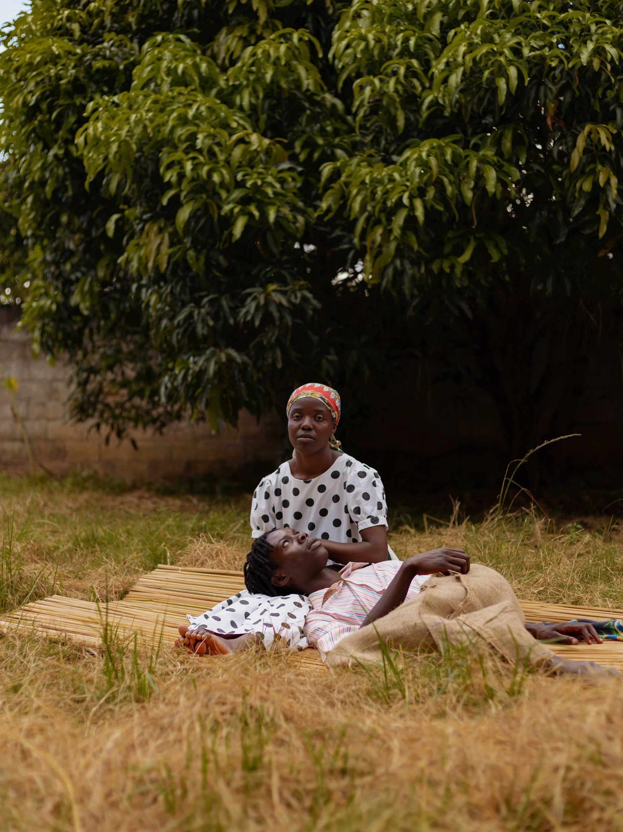 Two women, one lying down and another sitting, on a mat in an outdoor grassy area near a large tree. The woman lying down appears to be resting while the other woman looks directly at the camera. The scene is set in a natural environment with a brick wall in the background.