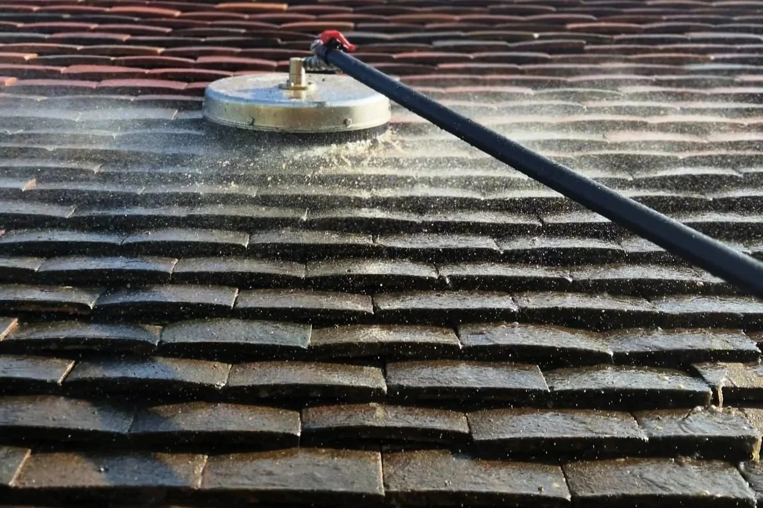 A tiled roof being cleaned with a professional pressure washer, with water spray visible on dark shingles.