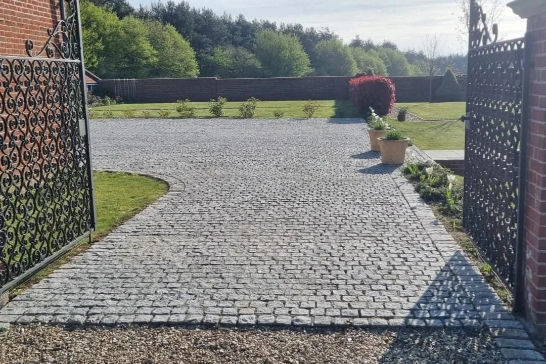View of a curved clean cobblestone driveway opening through black wrought iron gates, with potted plants along the edge and a green lawn with a red bush in the background.