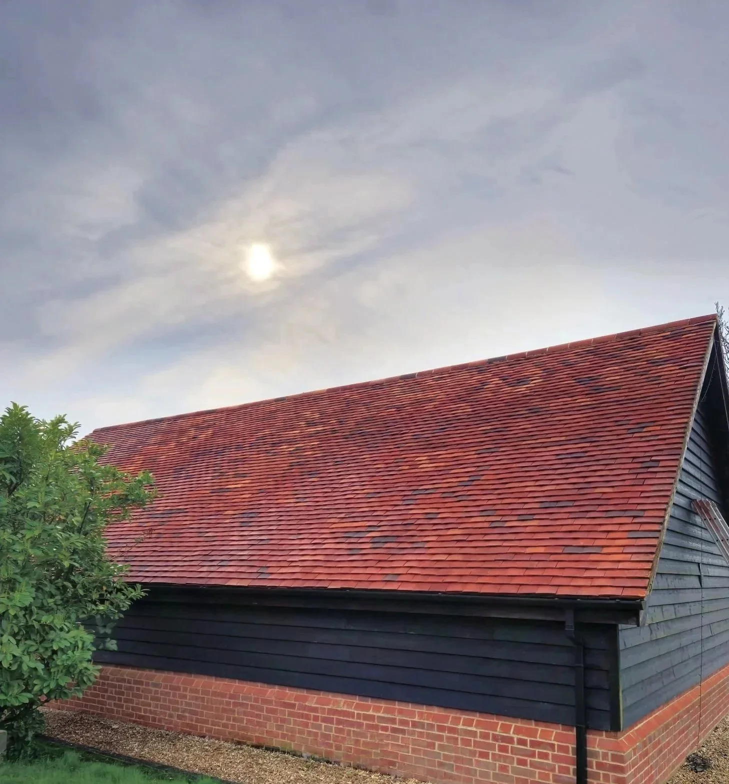A house with a freshly cleaned red tiled roof, dark wooden exterior walls, and a brick foundation, with some green foliage in the foreground and a cloudy sky with the sun partially obscured in the background.