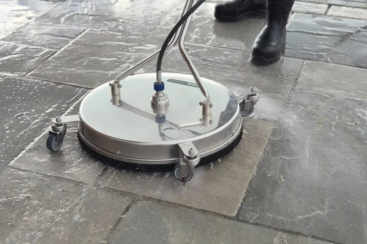 A professional patio and driveway floor cleaning tool on a stone tile floor driveway, with a person wearing black boots standing nearby.