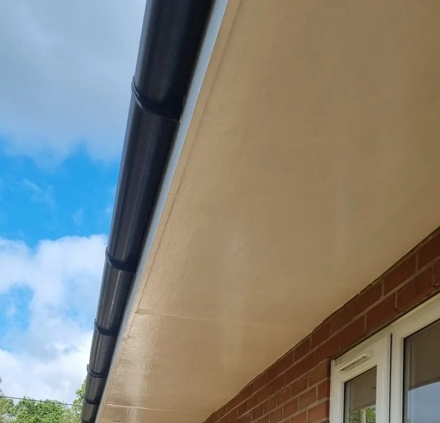 Close-up view of a house roof with black gutter, beige soffit, brick wall and window, under a partly cloudy sky.
