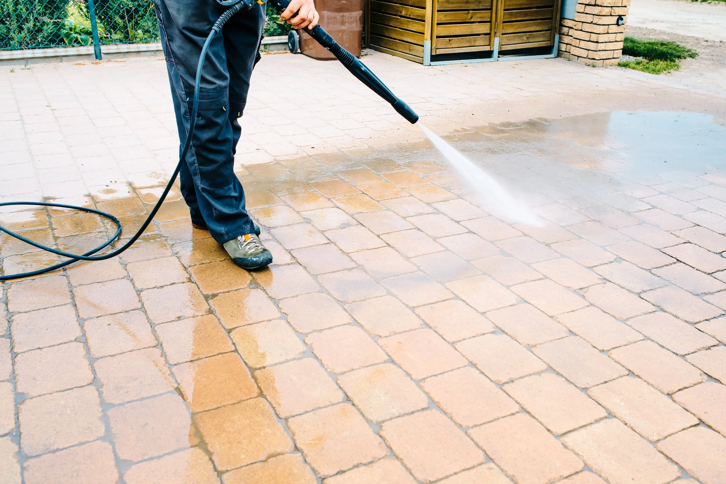 A professional exterior cleaning person wearing black trousers and gray sneakers is pressure washing a brick patio, spraying water on the surface.