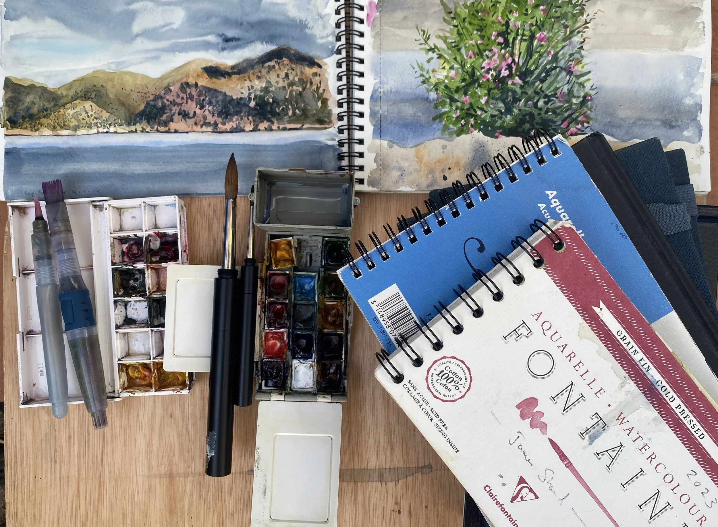 Une synthèse de matériel d'aquarelle comprenant des pinceaux, une palette de peintures, un carnet de croquis, et deux livres. En arrière-plan, il y a une peinture représentant une scène de montagne avec un lac et un ciel nuageux, ainsi qu'une peinture représentant un arbre en fleurs pink dans un paysage.