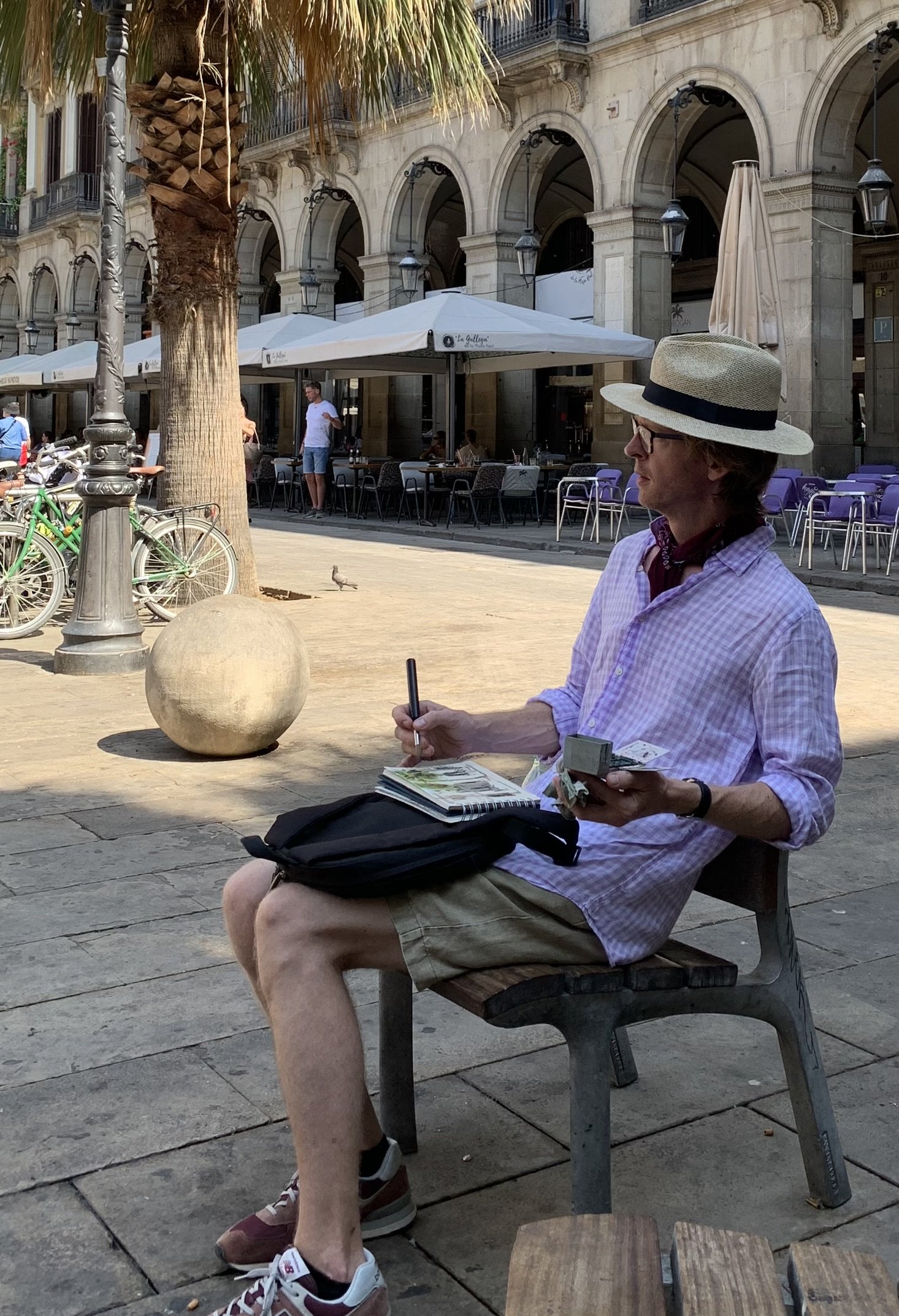 A man with a straw hat, glasses, a purple checkered shirt, and beige shorts sitting on a bench in a European city square, sketching in a notebook with a pen, holding some money and a small box, with outdoor cafes, bicycles, trees, and historic buildings in the background.