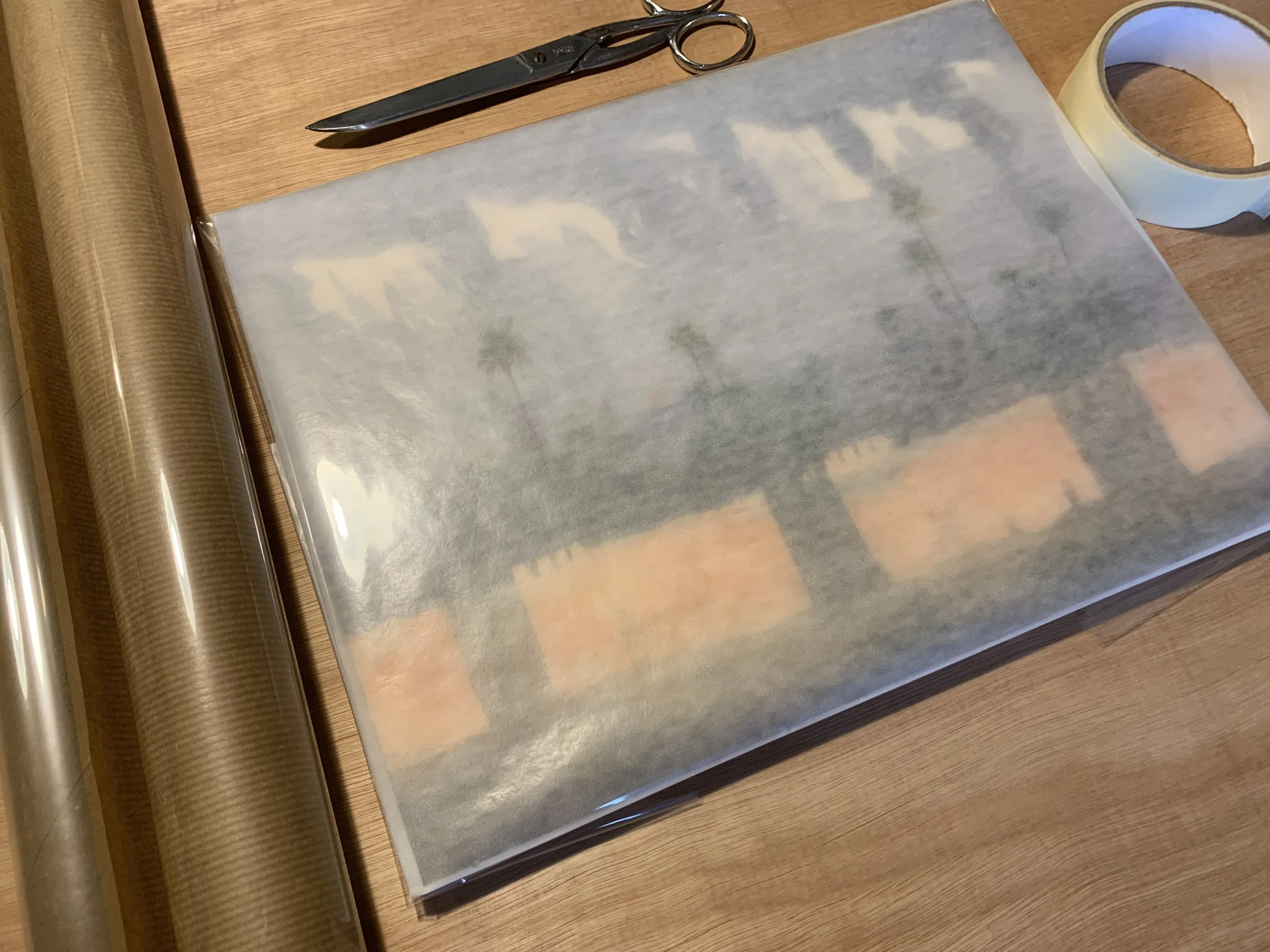 A sheet of wallpaper paper with a semi-transparent print of a house with a roof and palm trees, on a wooden surface. Surrounding items include scissors, painting tape, and a roll of clear protective film.