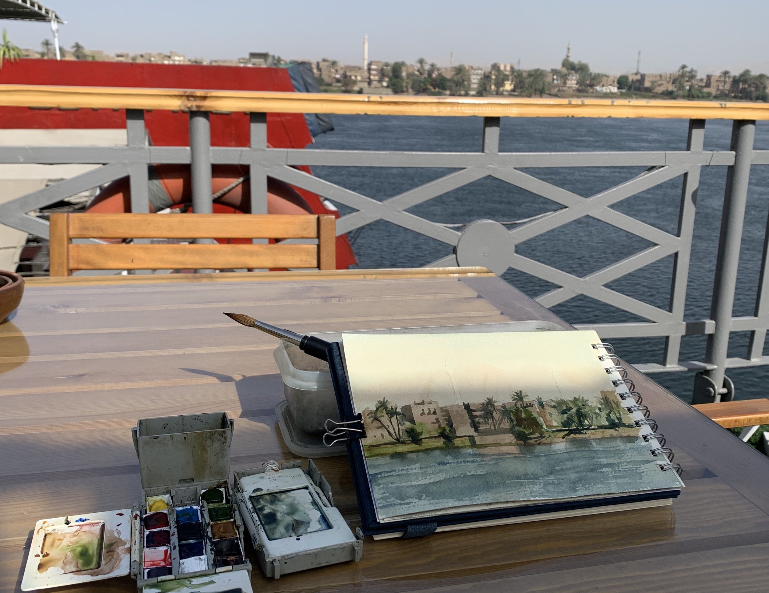 Watercolor painting supplies on a wooden table with a view of a river and cityscape in the background, including a sketchbook with a painting of palm trees and water, watercolor palette, and paintbrush.