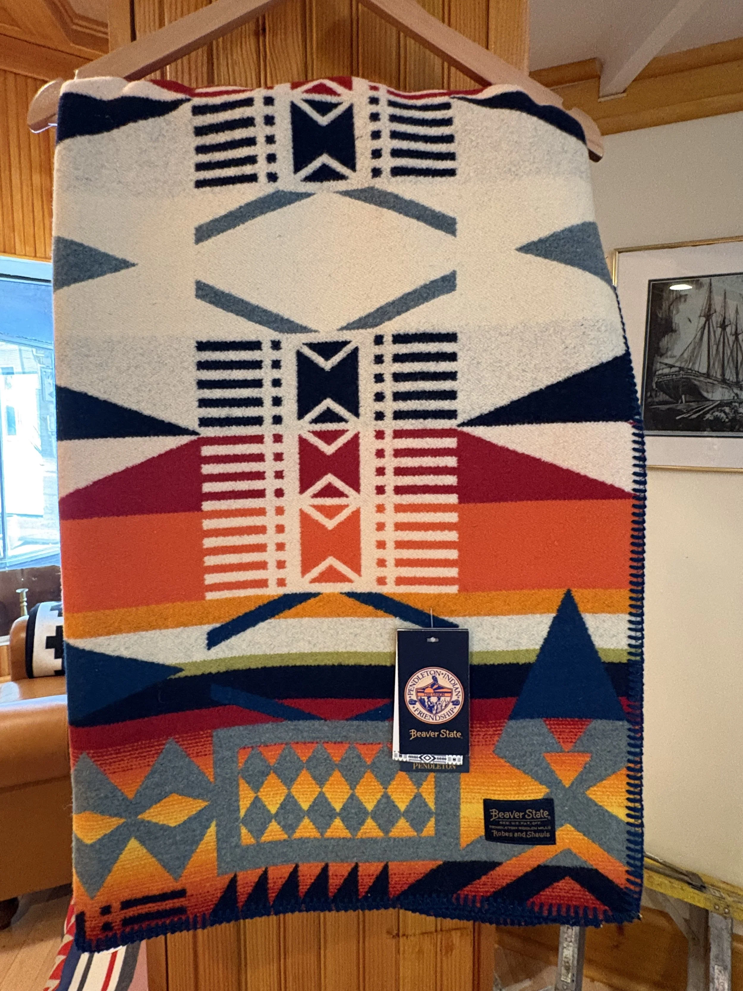 Colorful woven blanket with geometric and Native American-inspired patterns in red, orange, yellow, blue, and gray, hanging on a wooden wall with artwork in the background.