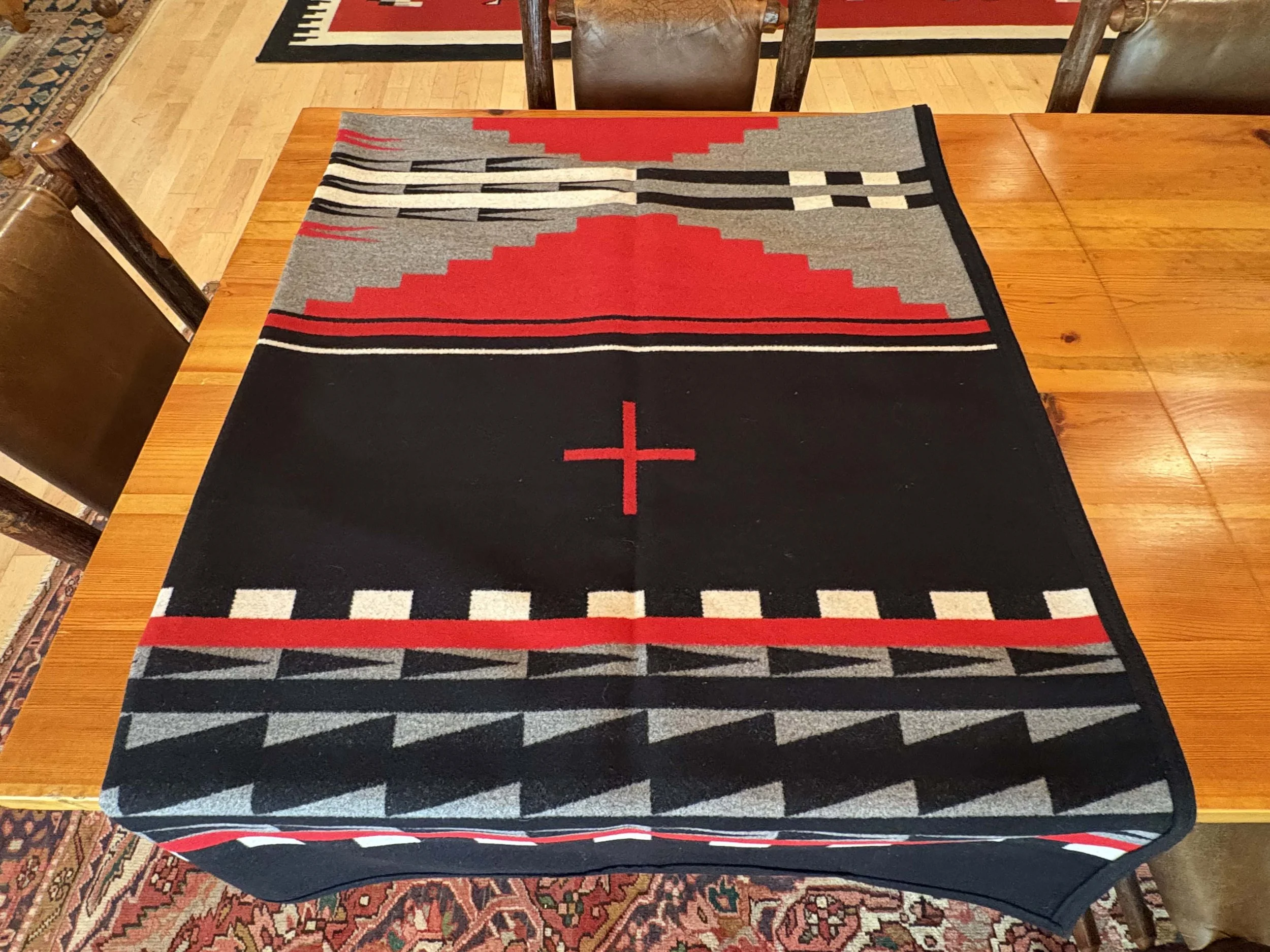 Table with a decorative woven table runner featuring geometric patterns in red, black, gray, and white. The surrounding area includes wooden chairs and a patterned rug.